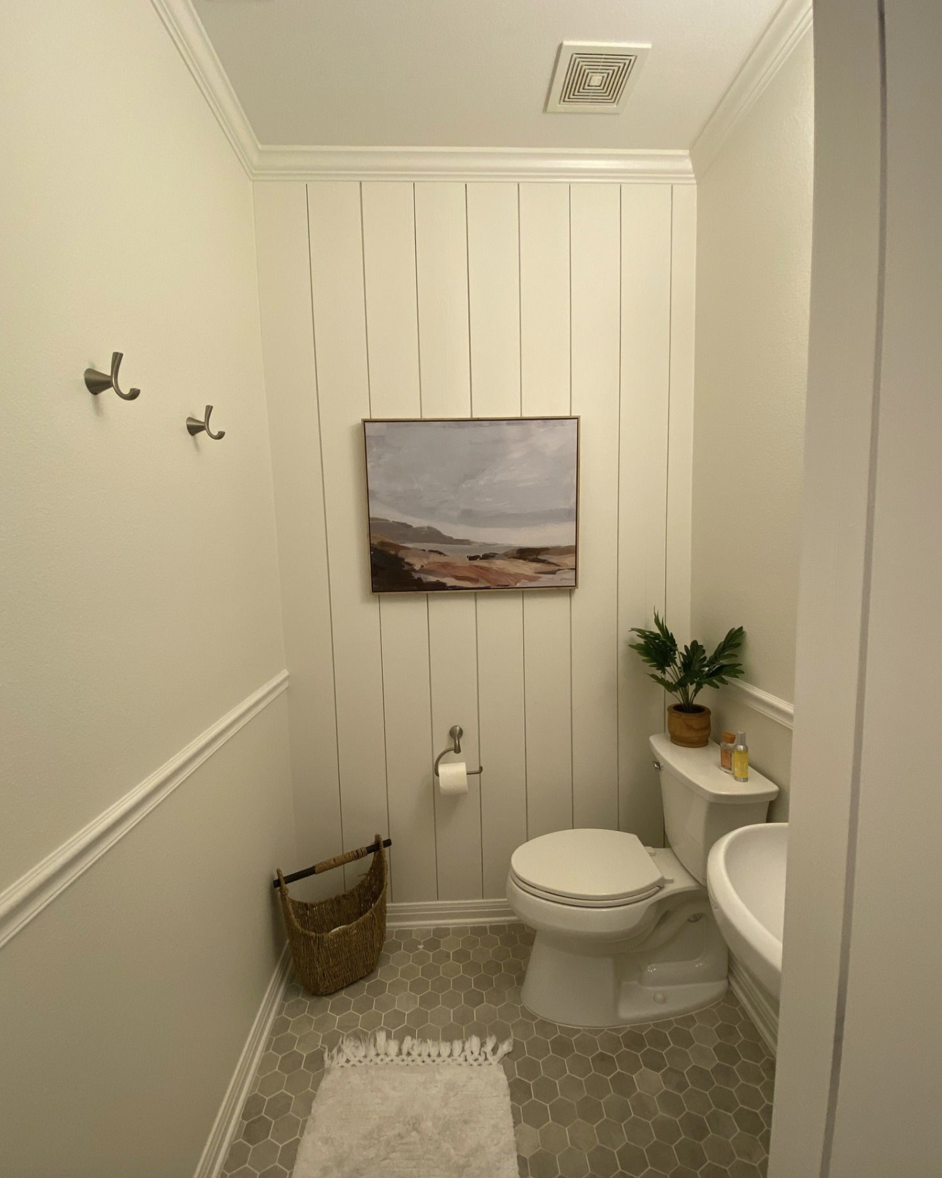 Small bathroom with light-colored walls and hexagonal tile flooring. A white toilet and sink are visible, with a small potted plant on top of the toilet tank. There are two hooks on the wall, a woven basket on the floor, a framed landscape painting on the wall, and a beige rug in front of the toilet.