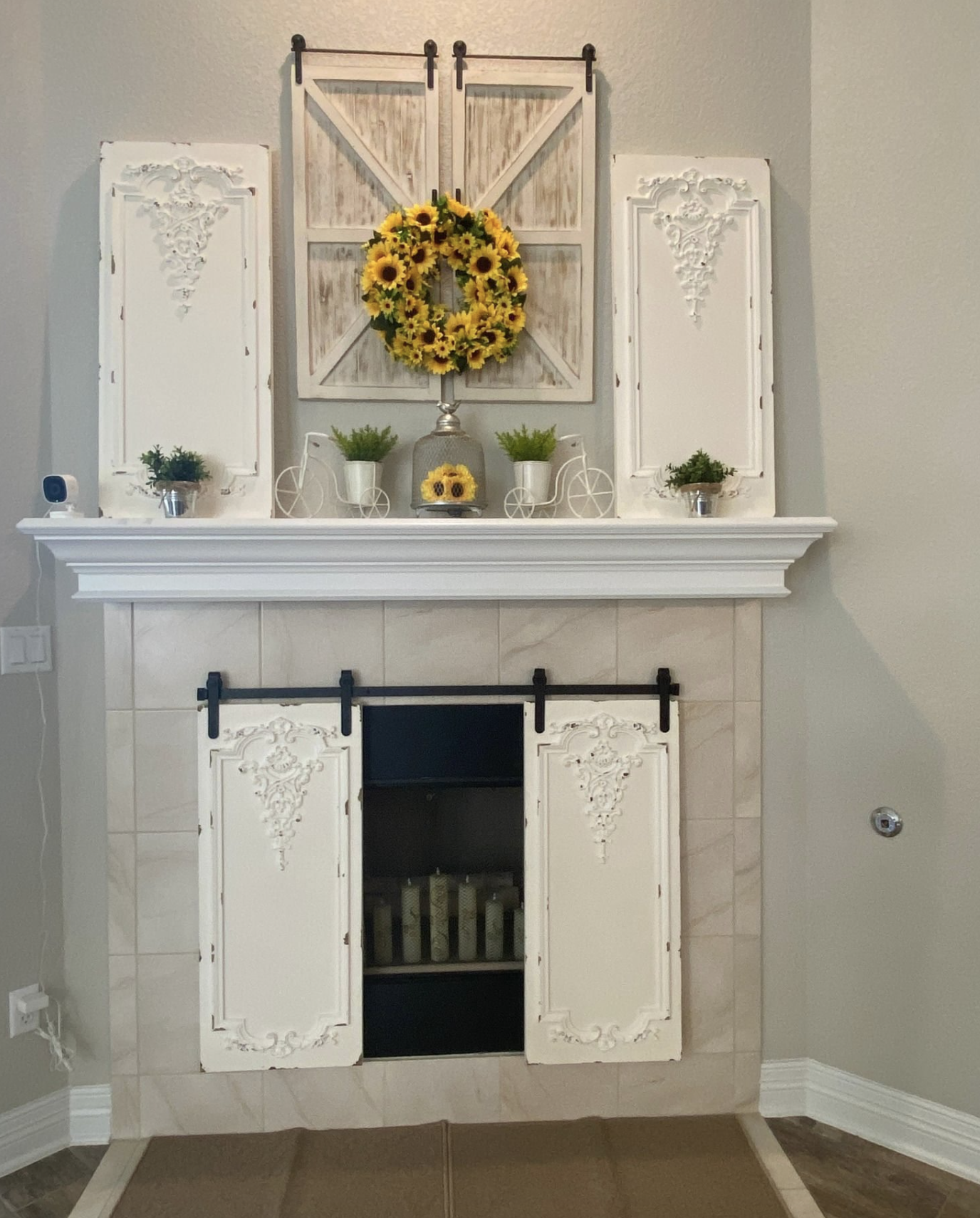 A white fireplace mantel with decorative panels, a black opening, and a tile surround. On top, there are small potted plants, miniature bicycles, and a sunflower wreath in the center. Above the mantel, a set of two rustic wooden barn doors with sunflower decorations is mounted.