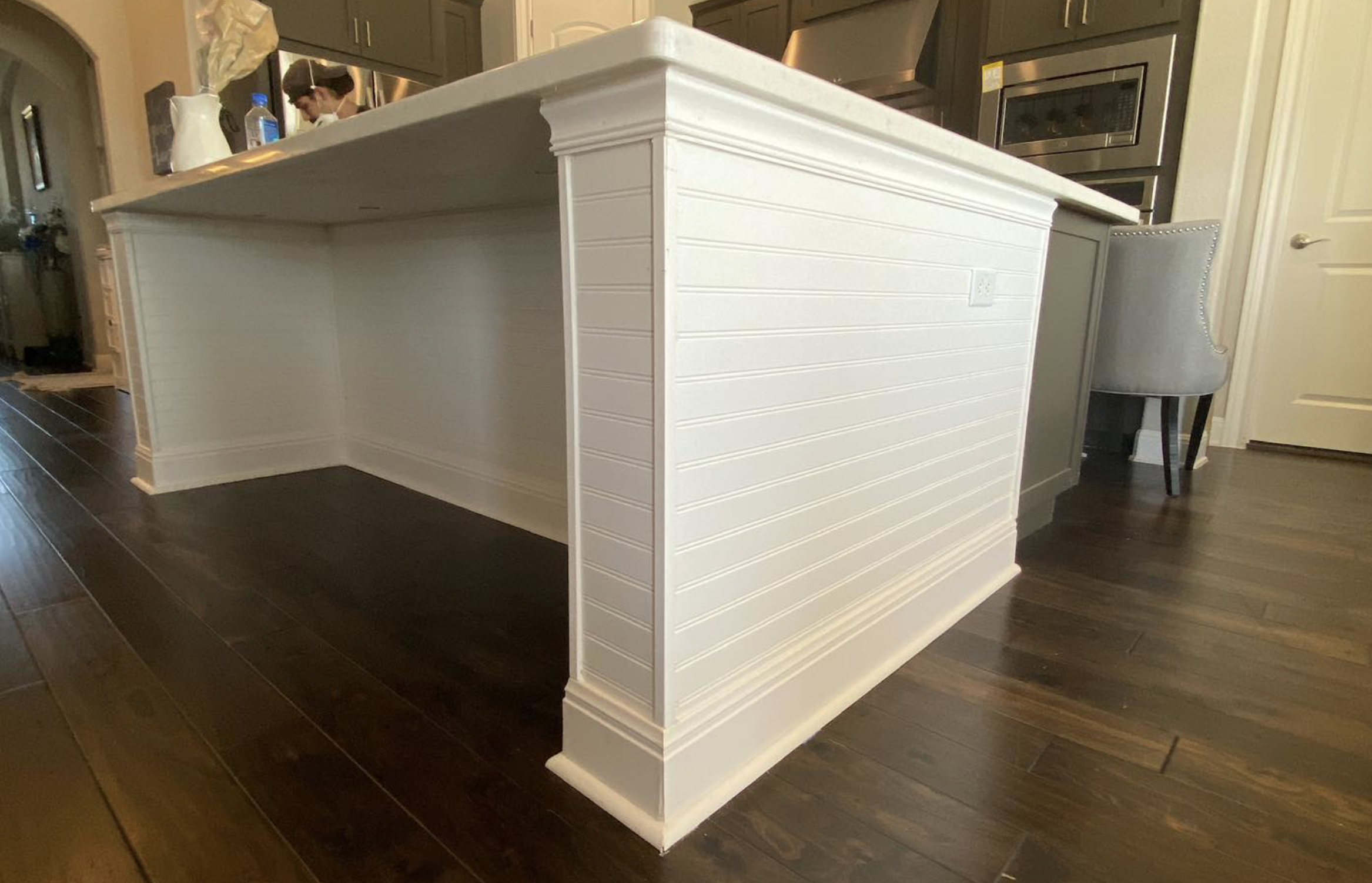 A kitchen island with white shiplap siding and a large white countertop, situated on dark hardwood floors. There is a woman in the background at the counter, with a metallic oven and a white door nearby.
