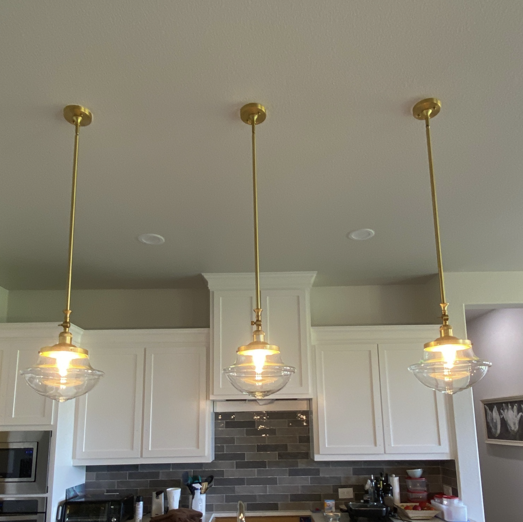 Three pendant lights with glass shades hanging from a ceiling above a kitchen counter with various items on it, including a microwave, a toaster oven, and kitchen utensils, in a kitchen with white cabinetry and a tiled backsplash.