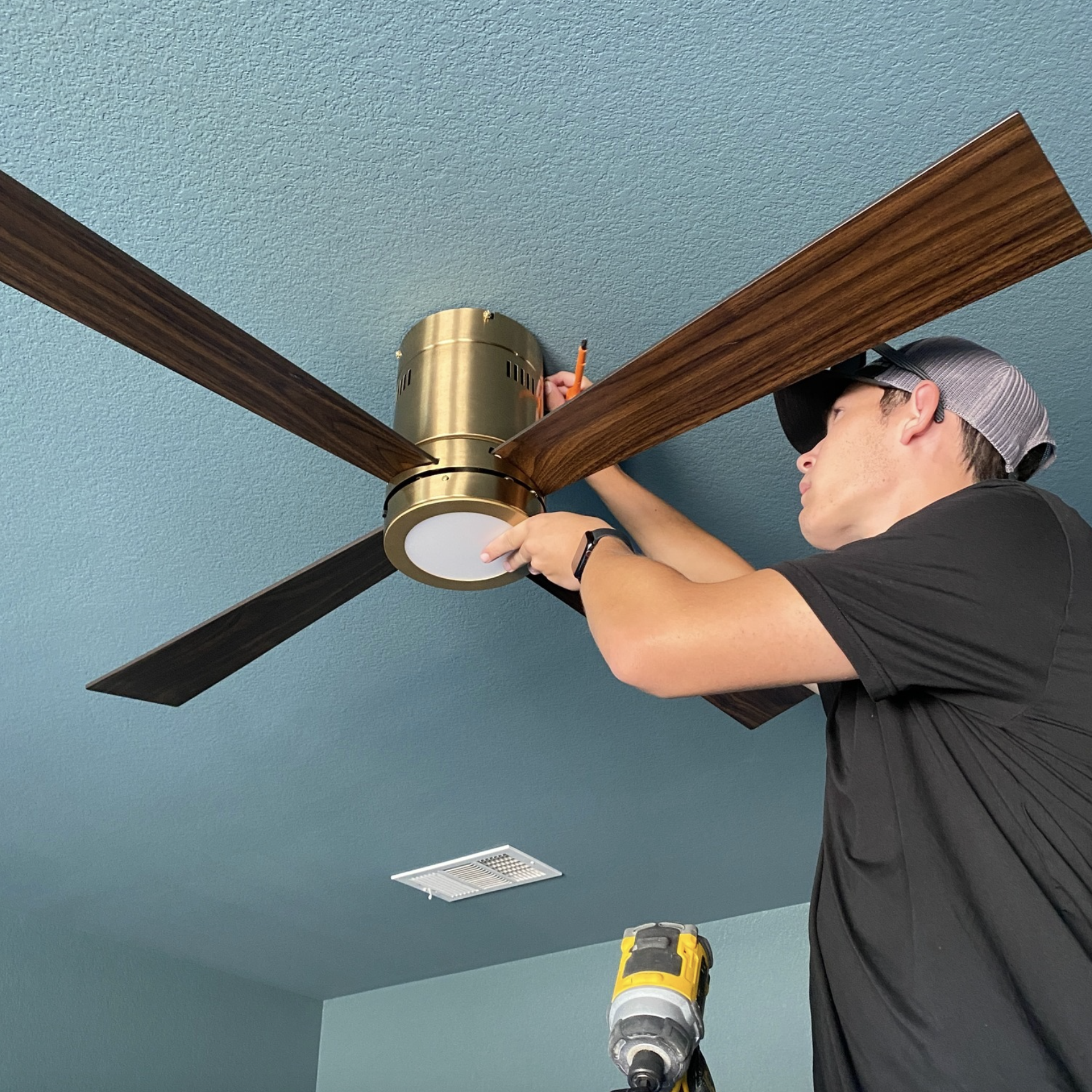 A man is installing a ceiling fan on a teal ceiling, using a cordless drill. He is wearing a baseball cap, black t-shirt, and a watch.