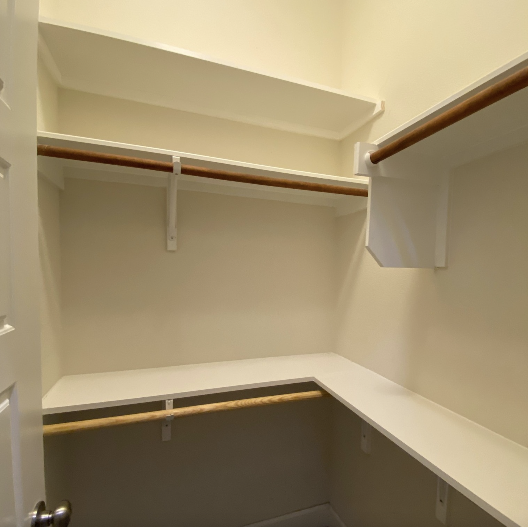 Empty walk-in closet with beige walls, wooden closet rods, and white shelving, viewed from open door.