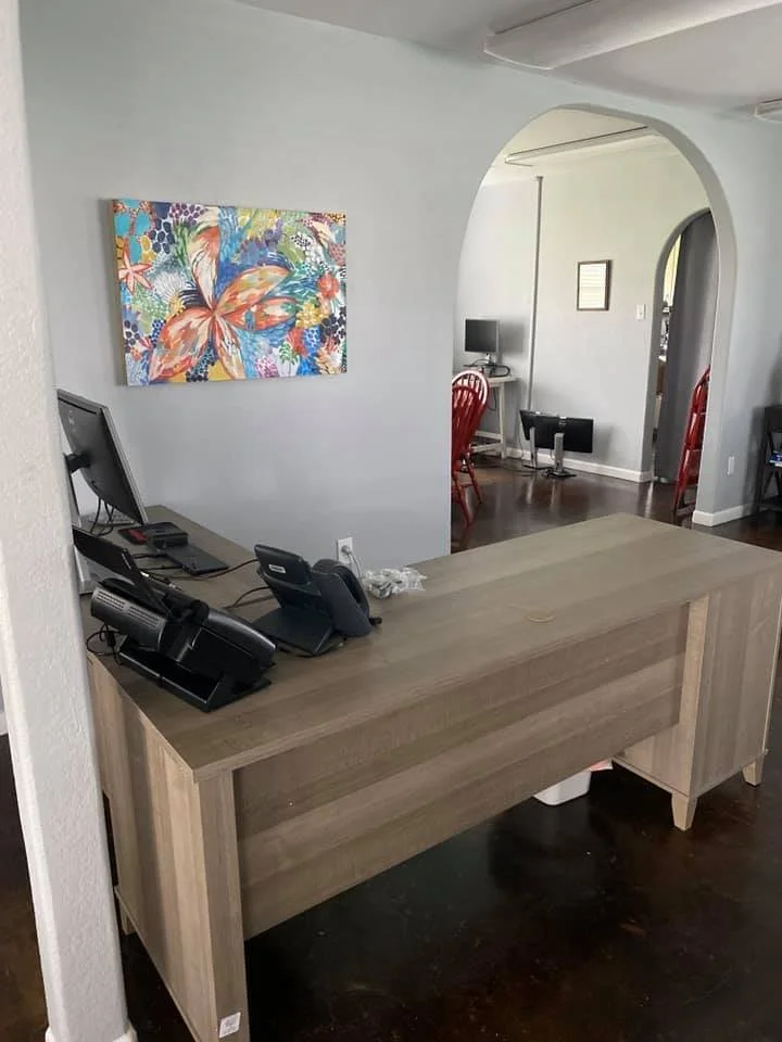 A wooden reception desk with a computer, telephone, and other office supplies, in a modern home office or reception area, with colorful artwork on the wall and a dining area with red chairs and a small desk with a computer in the background.