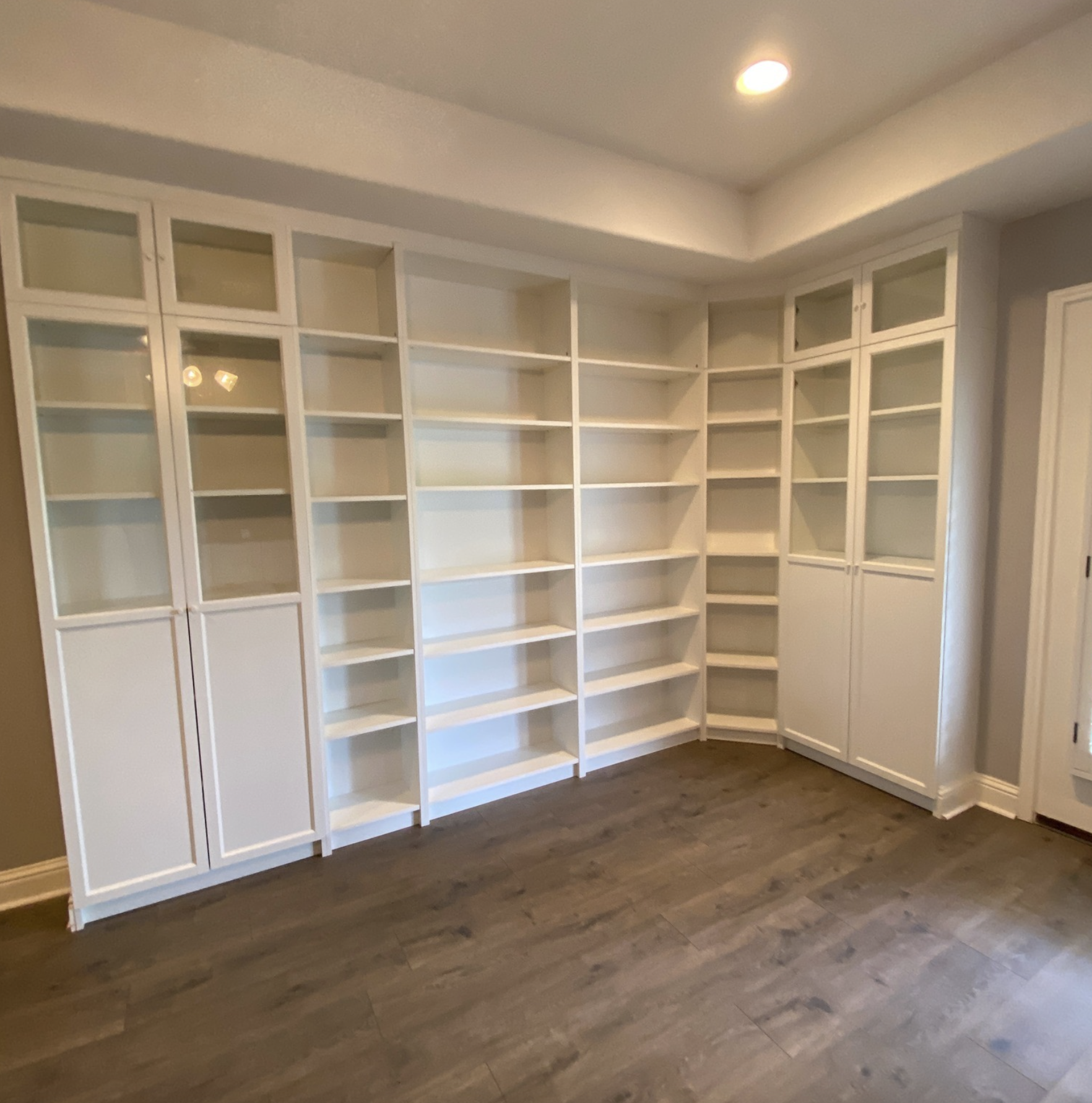 Empty white built-in shelves with glass-front cabinets, in a room with dark wood flooring and recessed ceiling lights.