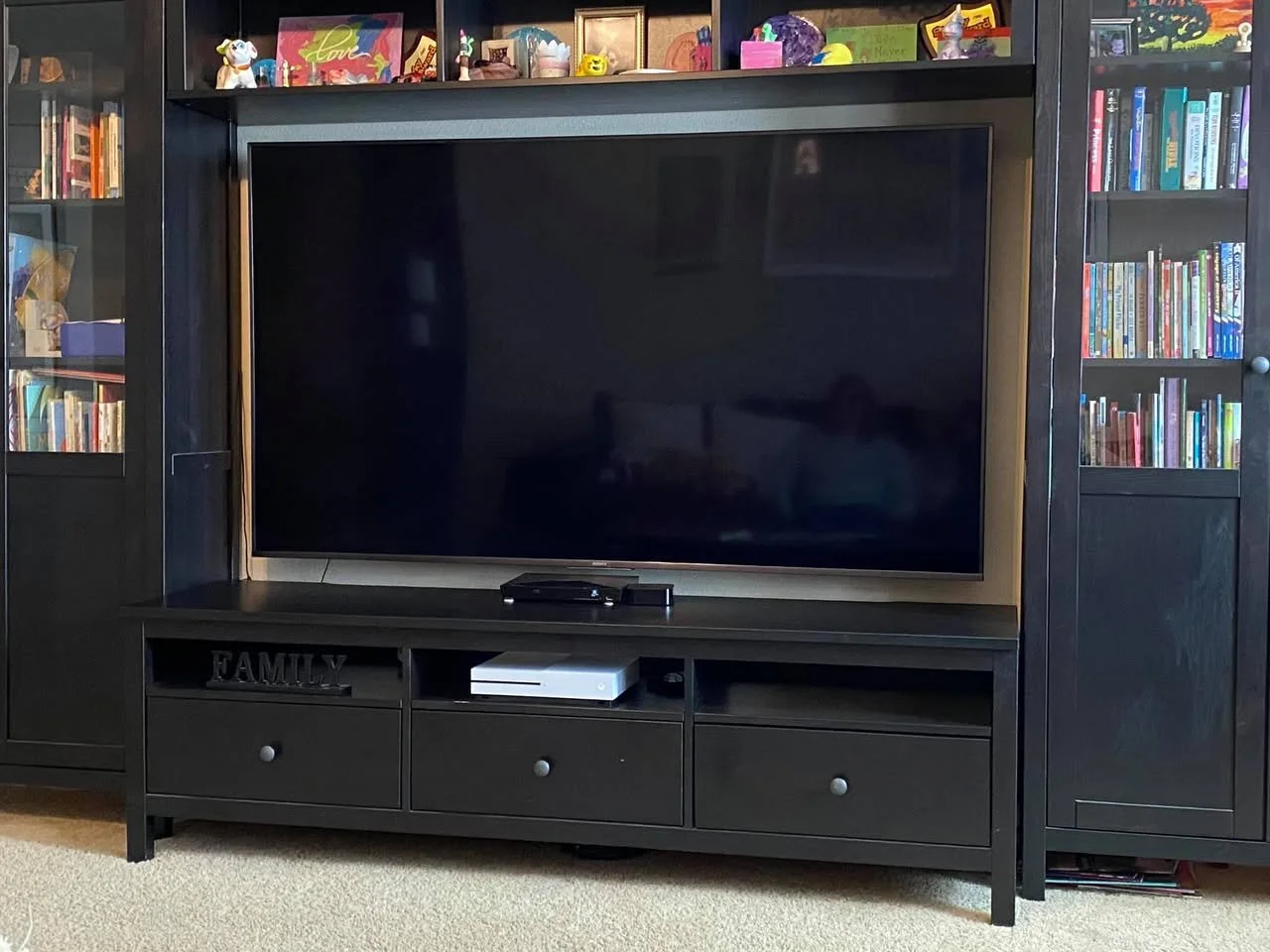 A black entertainment center with a large flat-screen TV, two glass-door bookshelves on each side filled with books, and a shelf above the TV holding various decorative items and picture frames.