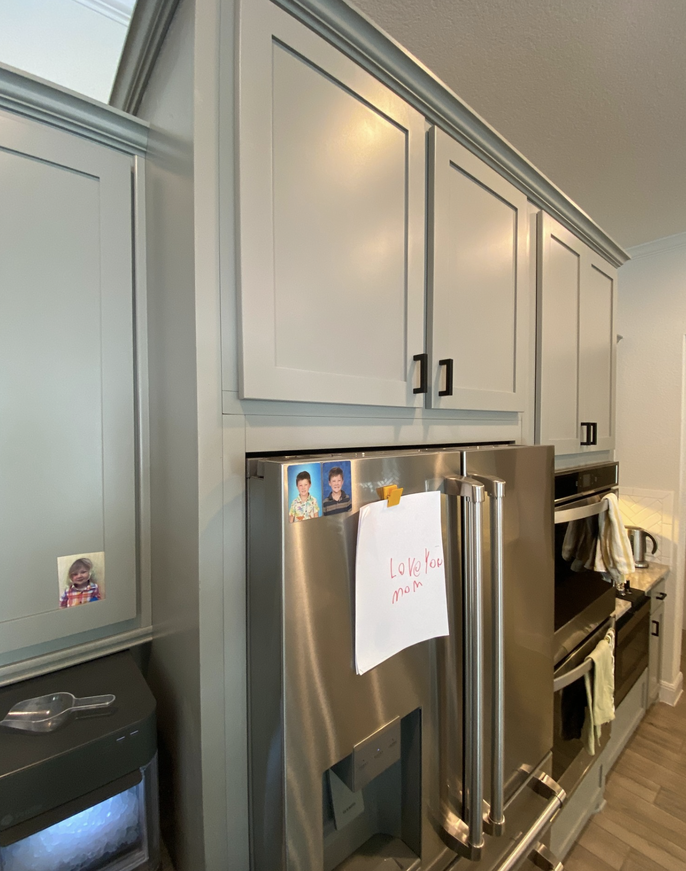Kitchen with light gray cabinets, stainless steel refrigerator with children's photos and a note that reads "Love you mom", and a black trash can. To the right, there is a stove with a towel hanging on the handle, and a countertop with a kettle and a