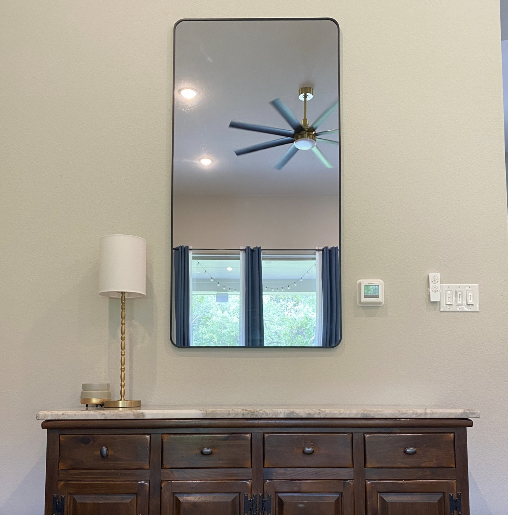 A mirror reflecting a ceiling fan and lights, a window with blue curtains, and a room outside with string lights. There is a white and gold table lamp on a wooden cabinet below the mirror and a wall with control switches and a thermostat.