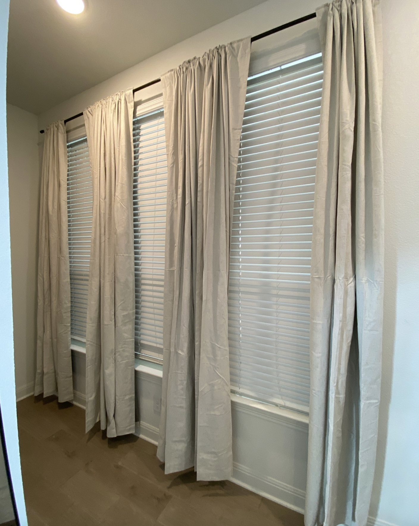 Four windows with white blinds and beige curtains hanging on black curtain rods in a room with wood flooring and white walls.