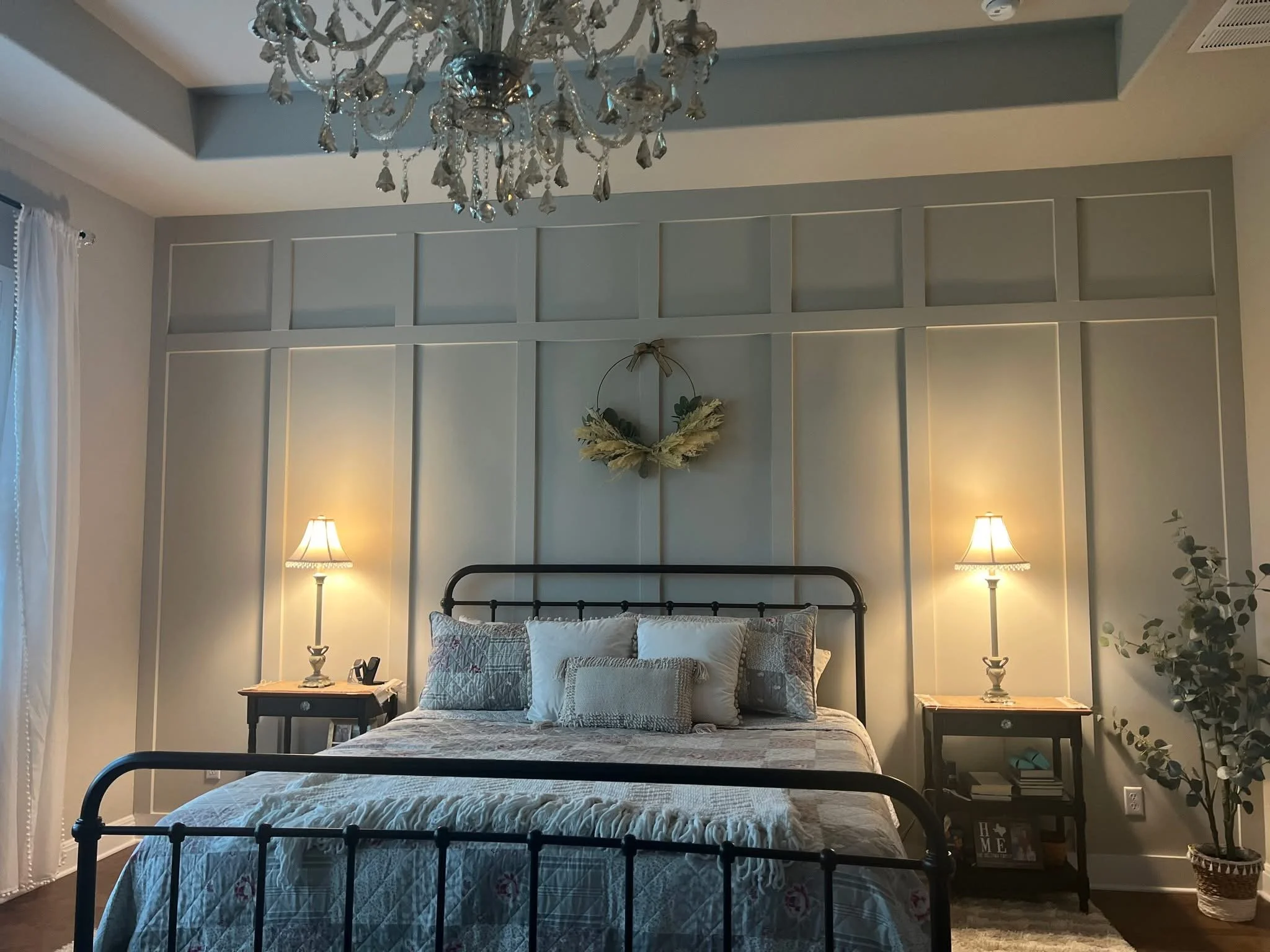 A bedroom with a black metal bed frame, a white quilt, and patterned pillows. Two nightstands with lamps, a wall with paneling and a wreath, a chandelier hanging from the ceiling, and sheer white curtains over a window.