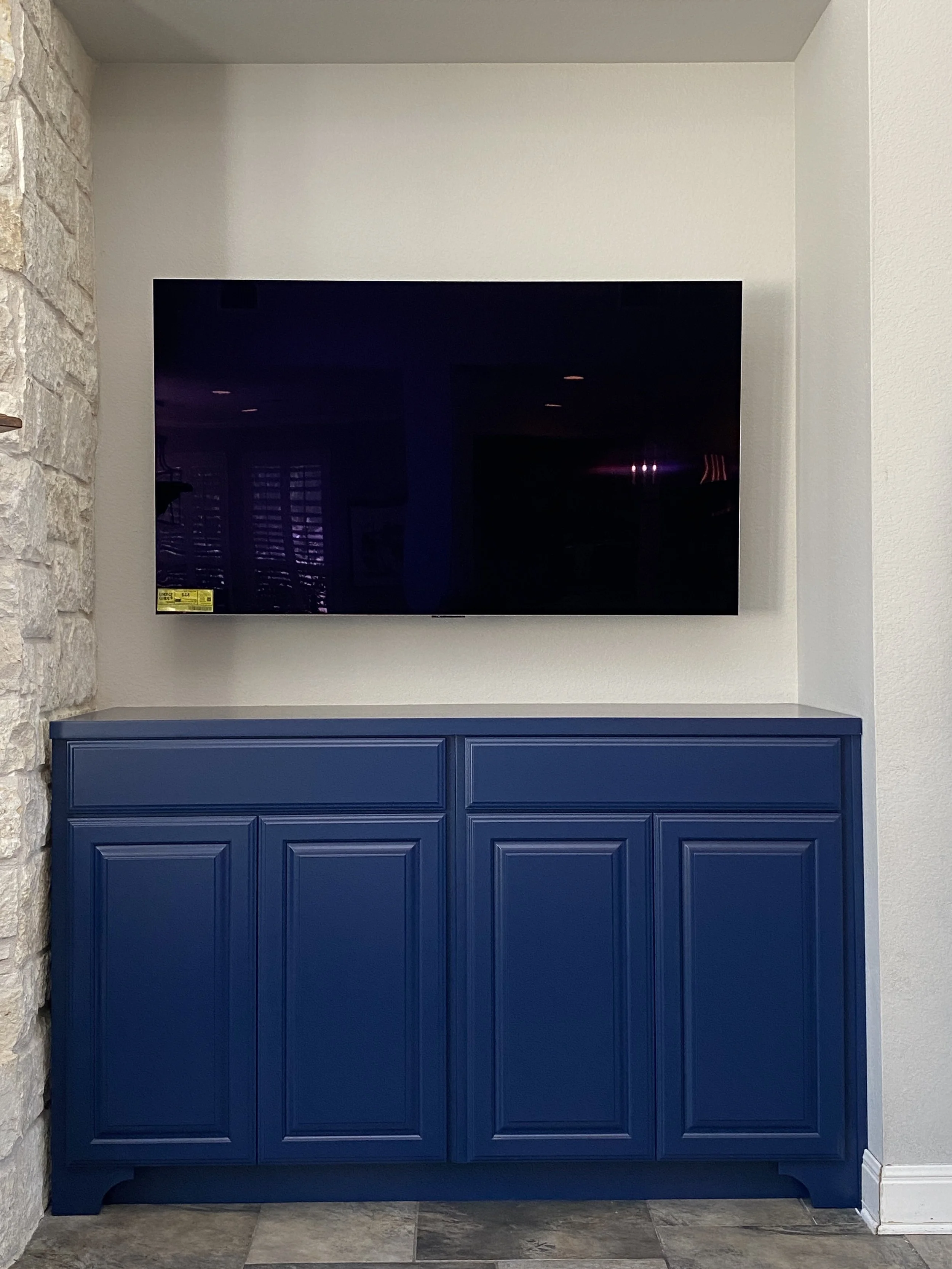 A flat-screen TV mounted on a white wall above a blue cabinet, with a stone wall on the left and a gray tiled floor.
