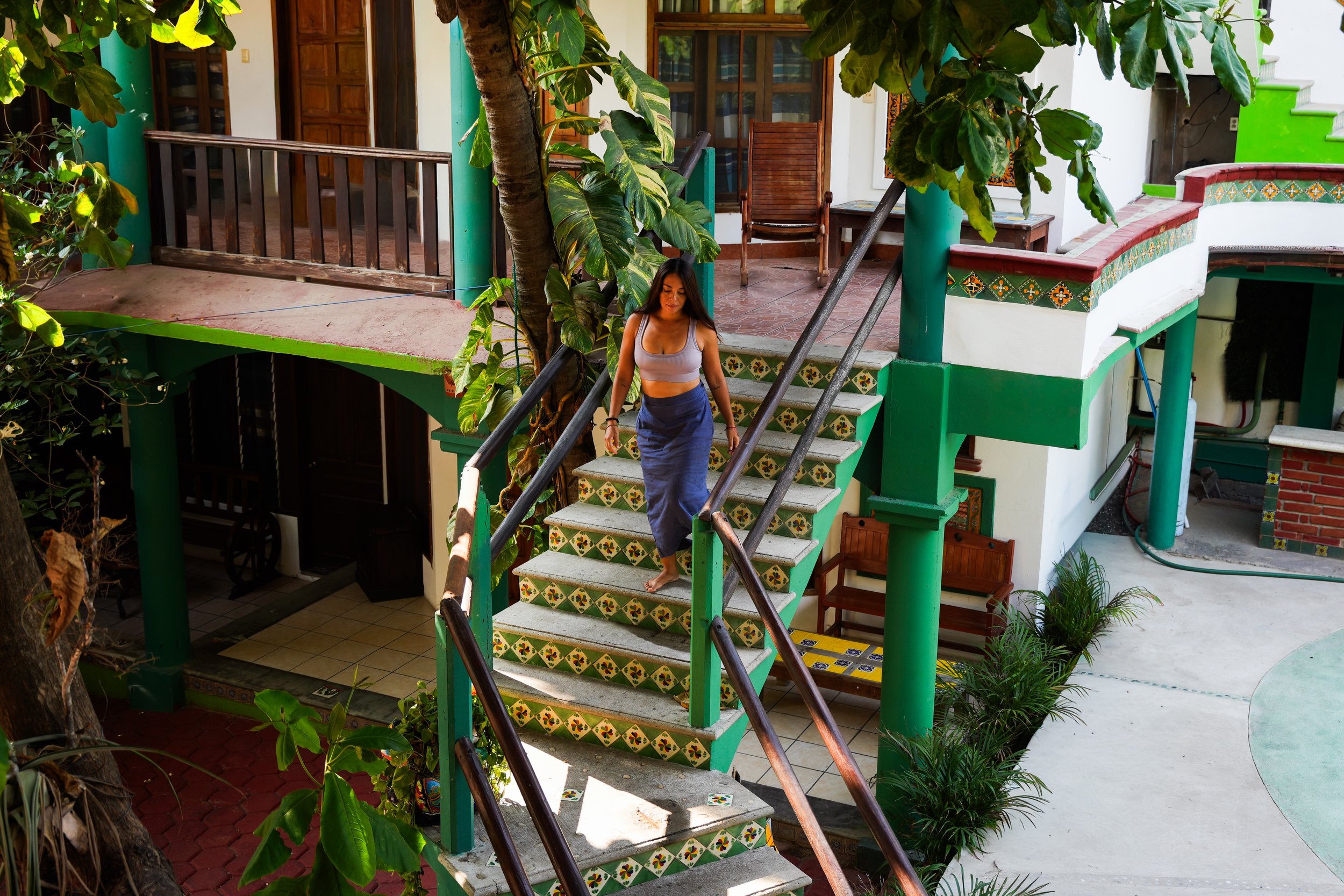A woman with tan skin, long dark hair, wearing a gray tank top and a blue skirt walking down colorful tiled outdoor stairs with green railings, surrounded by plants and trees, in front of a white house with wooden doors and windows.