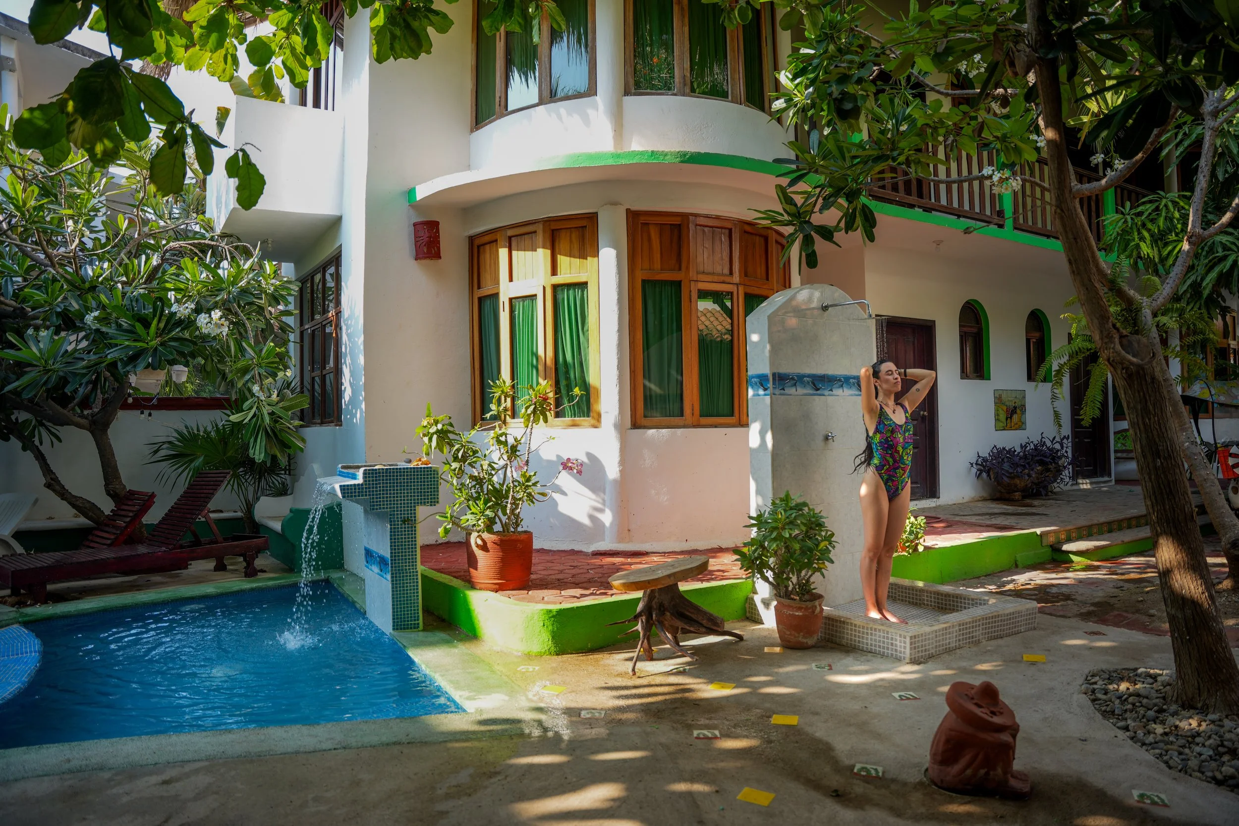 A woman in a colorful swimsuit standing near a pool in a lush garden yard, surrounded by trees and plants, with a white house featuring large windows and wooden accents in the background.