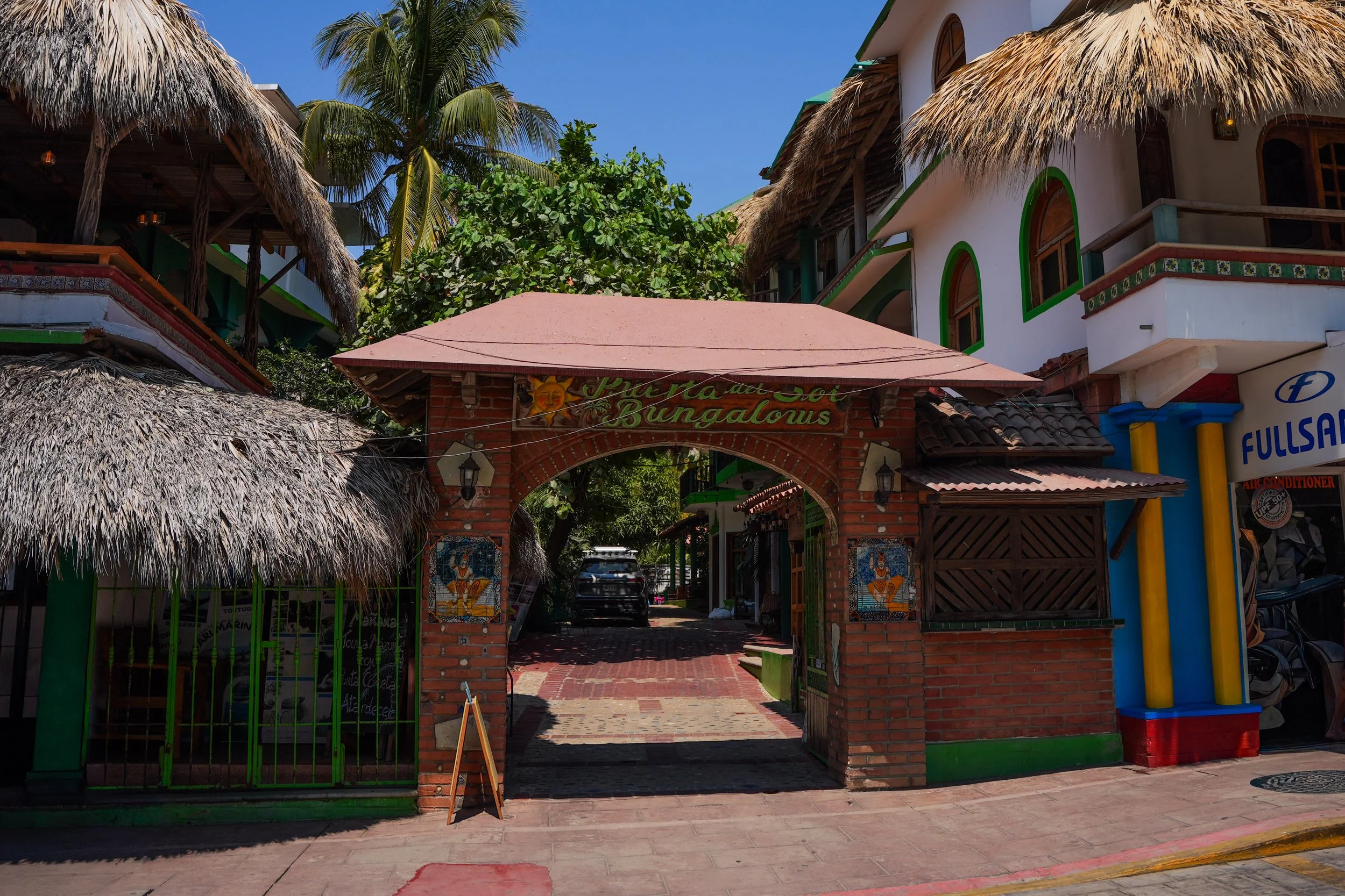 Entrance to a building with a sign that says 'Casa del Sol Bungalows,' featuring a brick archway, tiled murals, and tropical thatched roofs with palm trees in the background.