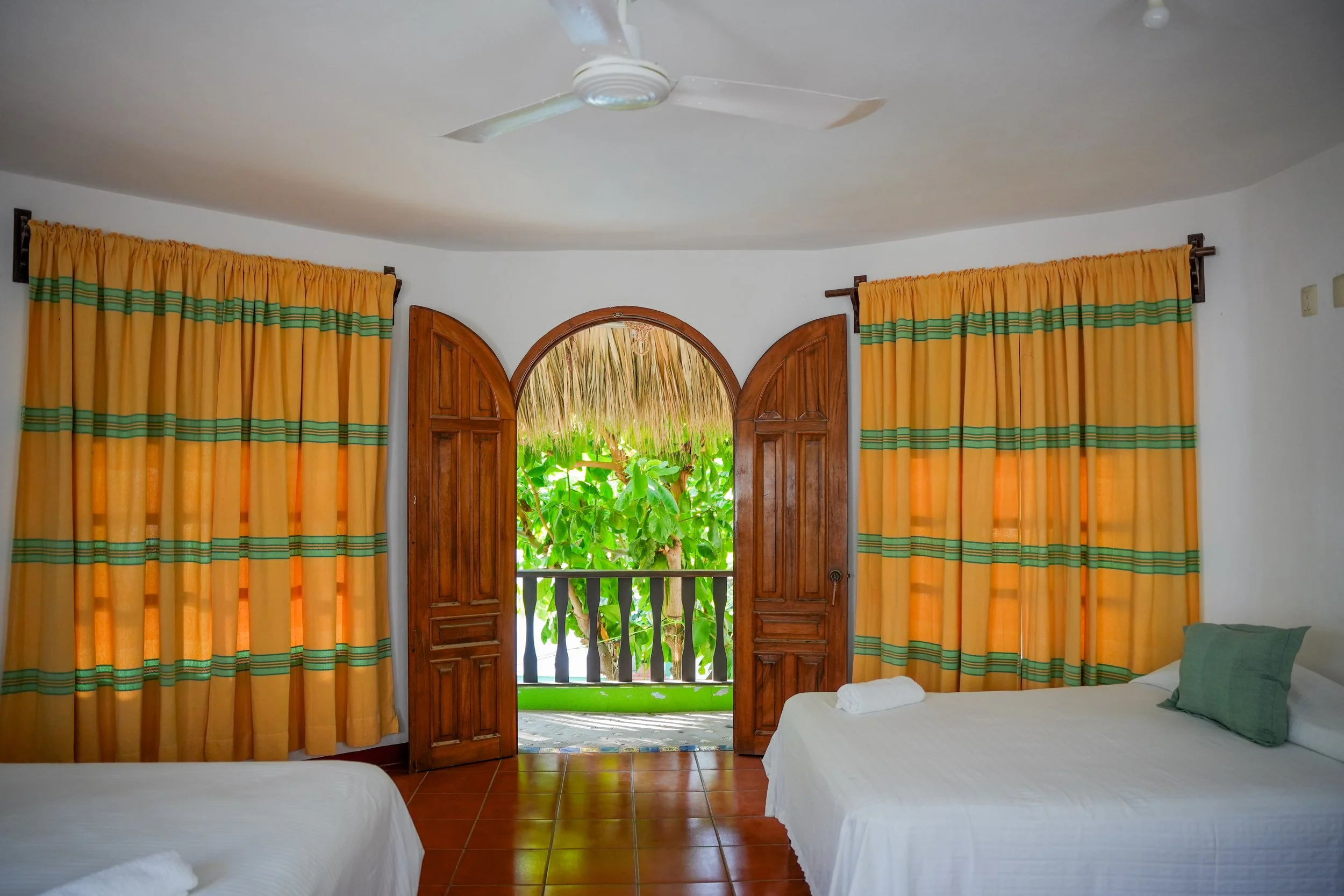 Room with white walls, yellow curtains with green stripes, wooden arched window showing green foliage outside, two beds with white linens and green pillows, and a ceiling fan.