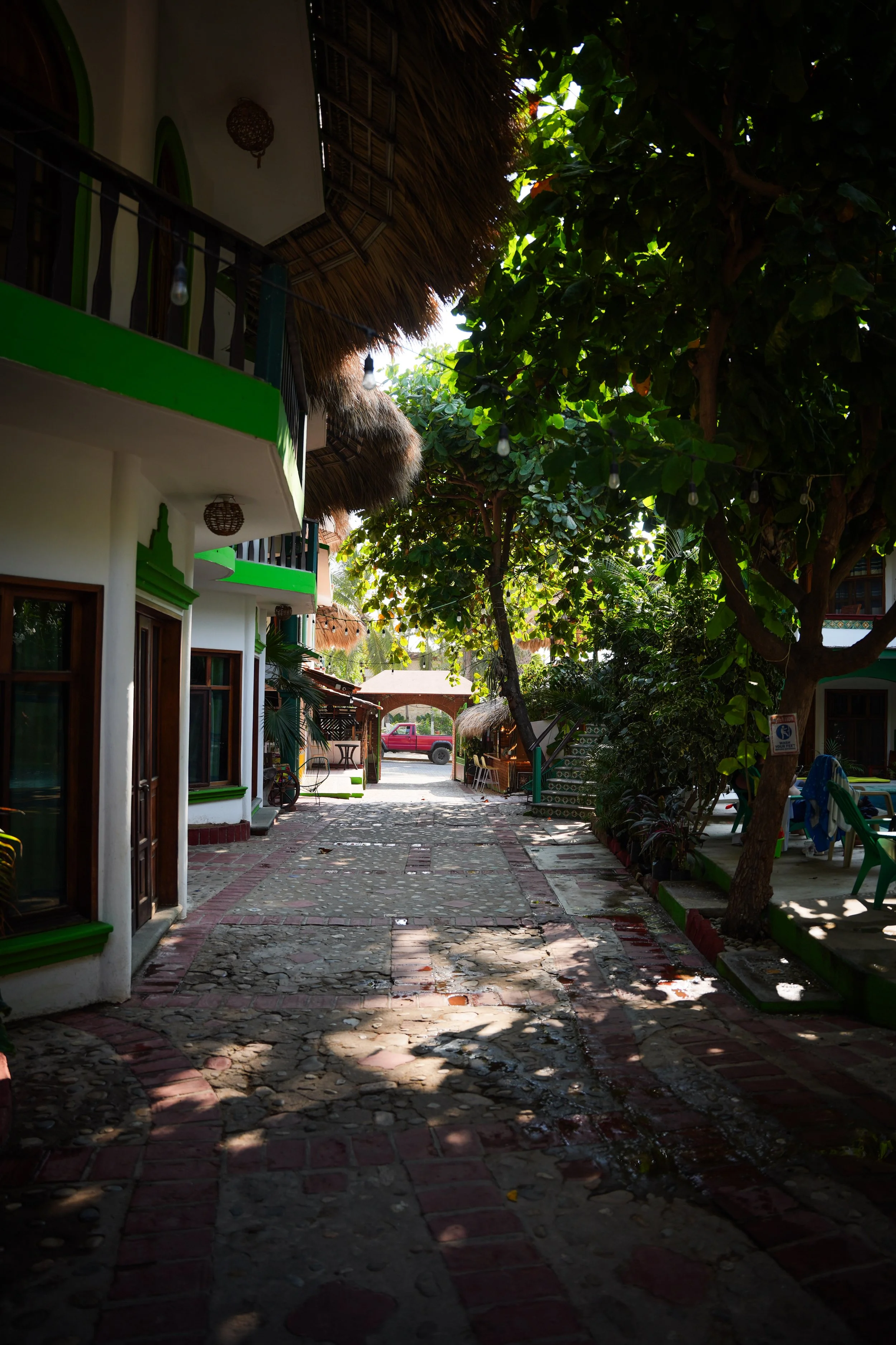 A cobblestone street with shaded areas and some wet patches, flanked by buildings with white walls and green accents, and lush green trees overhead providing partial shade.