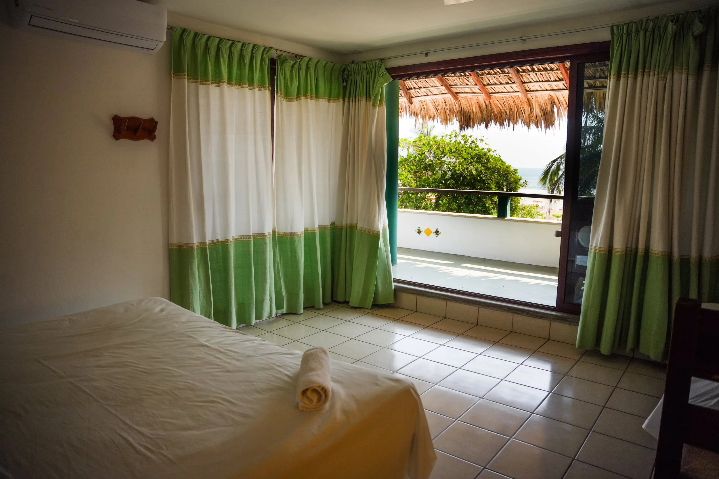 A hotel room with a bed covered in a beige bedspread, a towel rolled at the foot, green and beige curtains, a balcony with a thatched roof, trees, and a partial view of the ocean in the background.