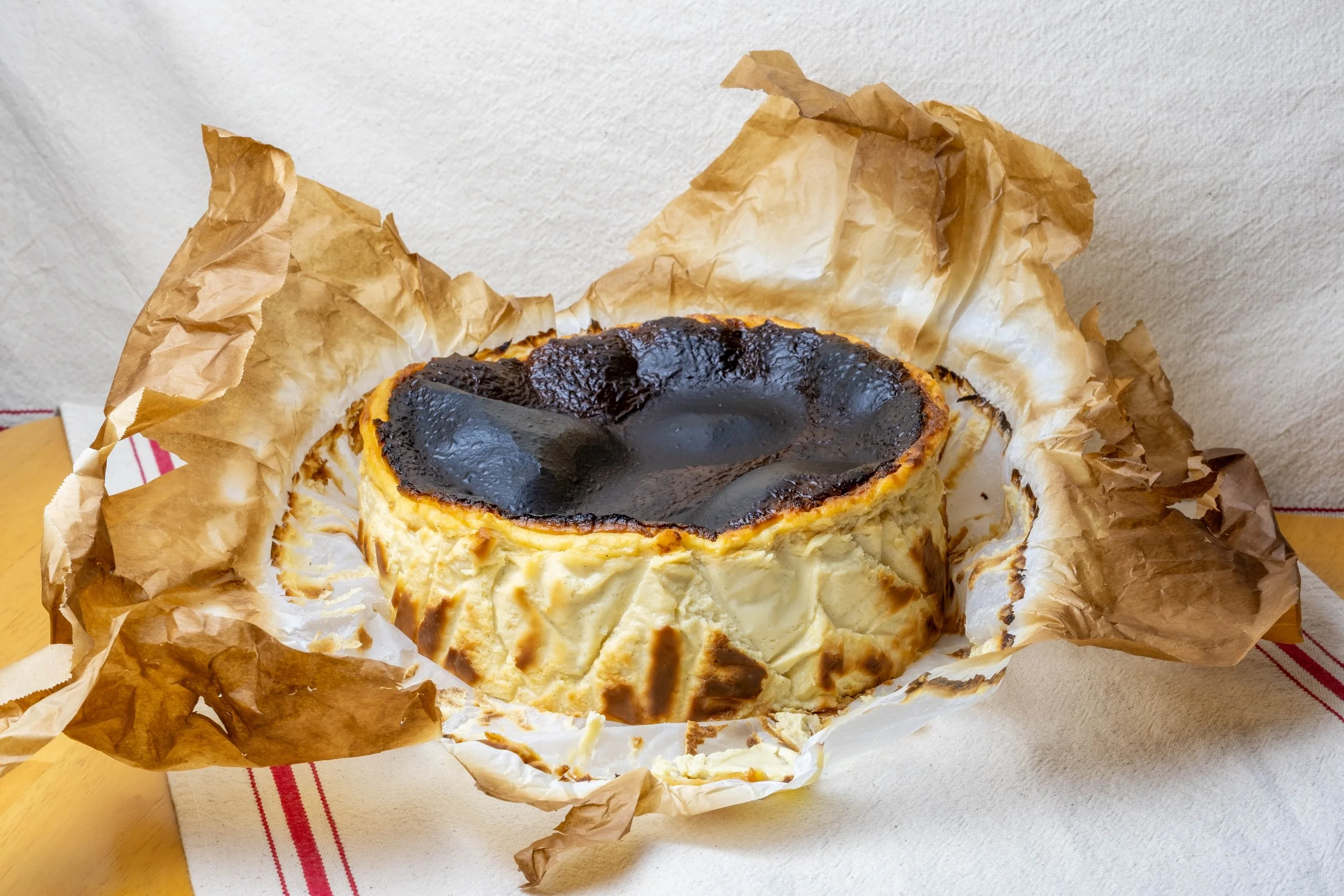 A burnt cheesecake wrapped in crumpled parchment paper, partially eaten, sitting on a white surface with a striped cloth underneath.