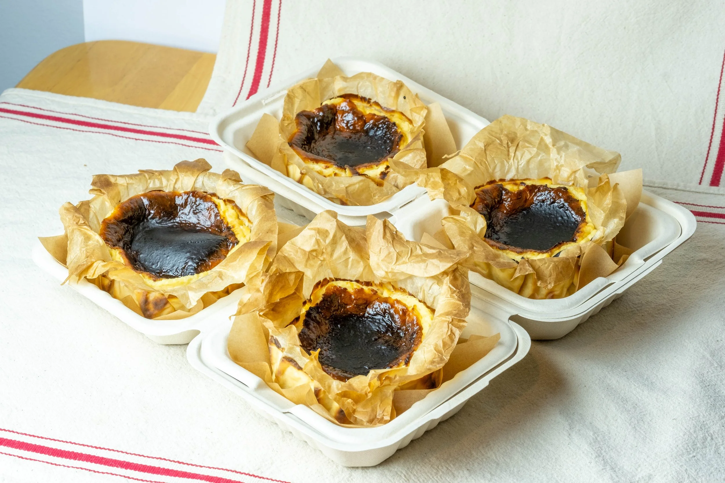 Four bento Basque cheesecakes with dark caramelized tops in white containers lined with crinkled parchment paper, placed on a white cloth with red stripe detail.