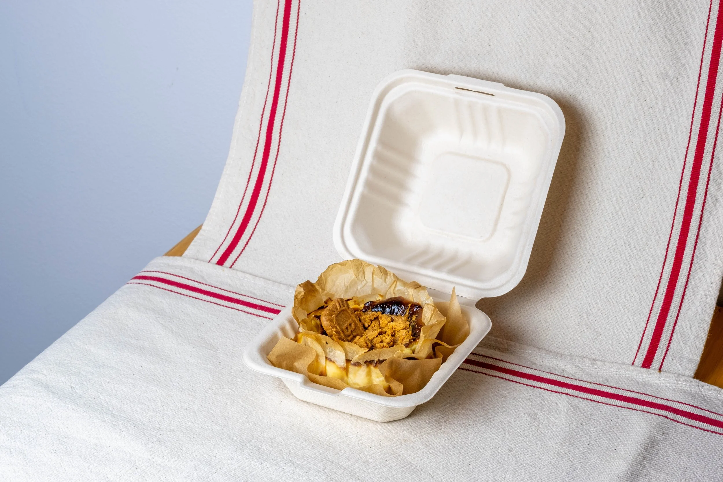Bento Biscoff Basque cheesecake topped with cookie butter in a to go container, placed on a white table with a red-striped cloth.