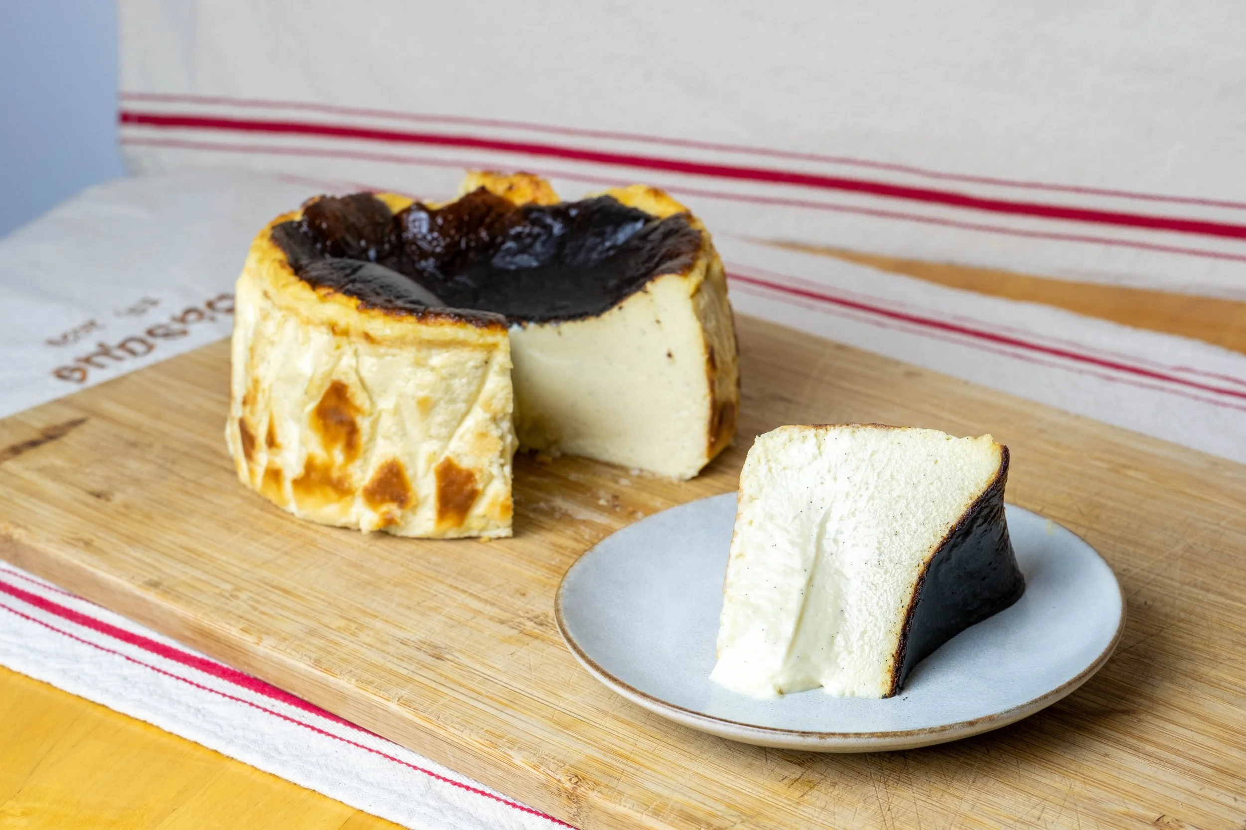 A round cheesecake with a dark chocolate top is on a wooden cutting board. A slice has been cut and is placed on a small plate nearby.