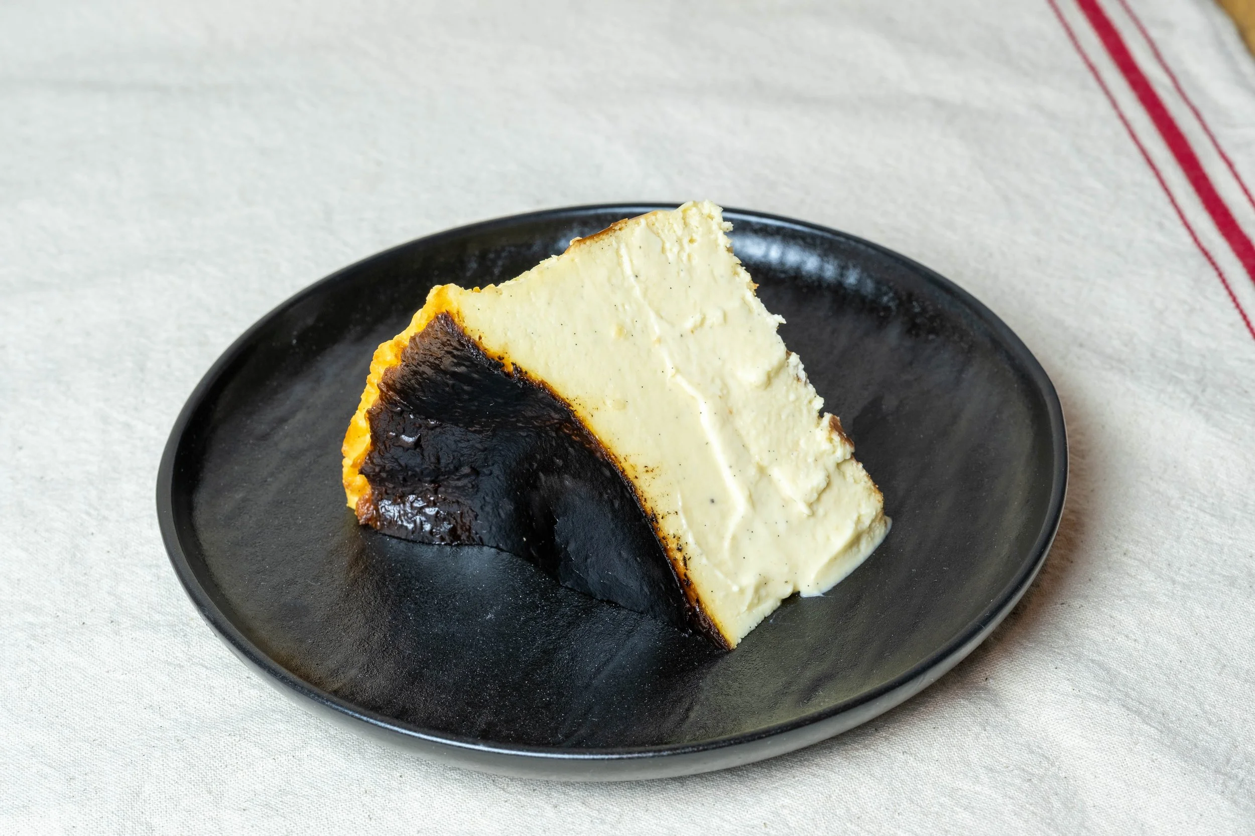 A slice of cheesecake with a dark chocolate crust on a black plate, placed on a light-colored fabric surface with red stripes.