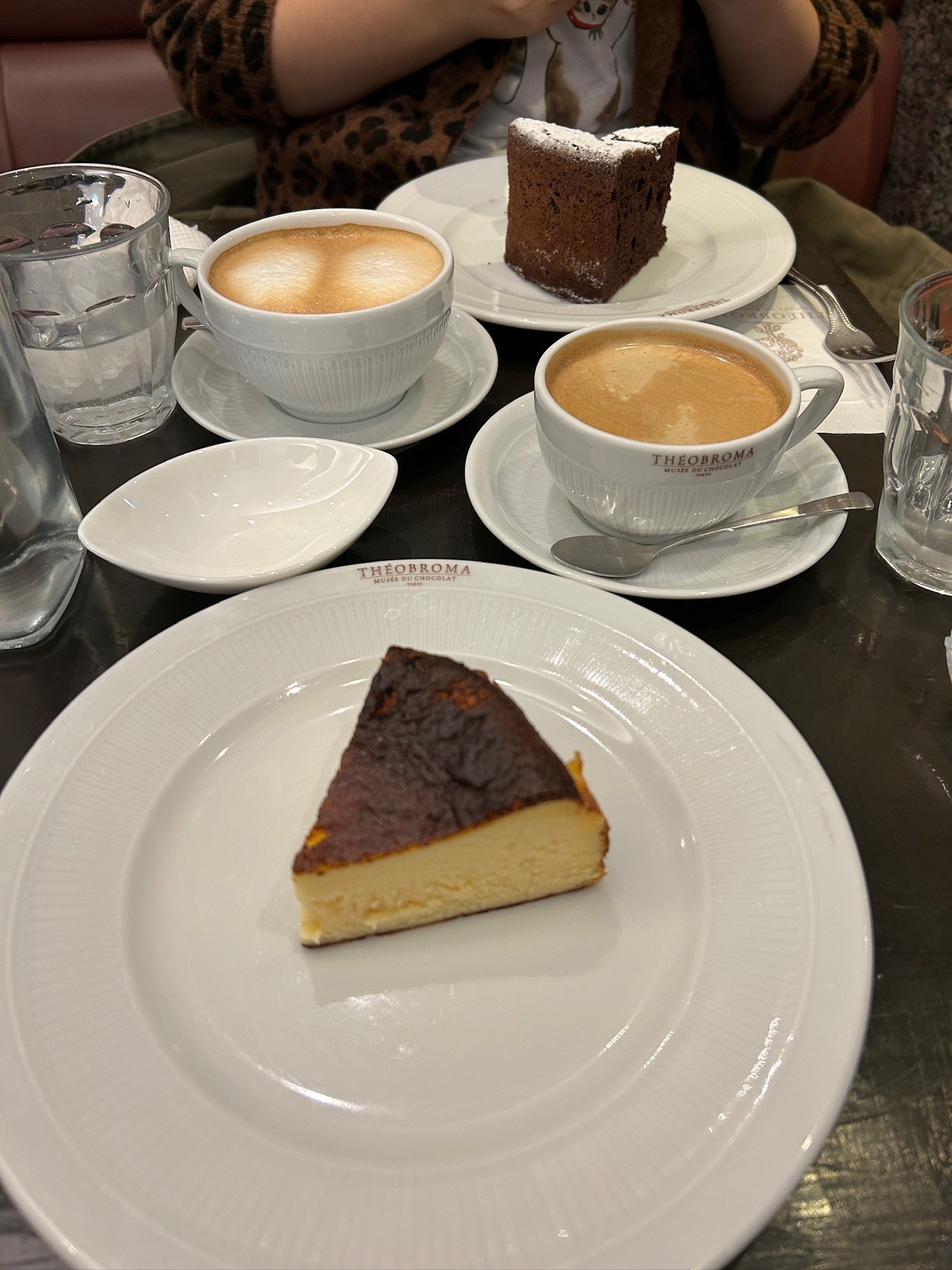 Slice of cheesecake on a white plate in the foreground, two cups of coffee with frothy milk on saucers, a piece of chocolate cake on a white plate in the background.