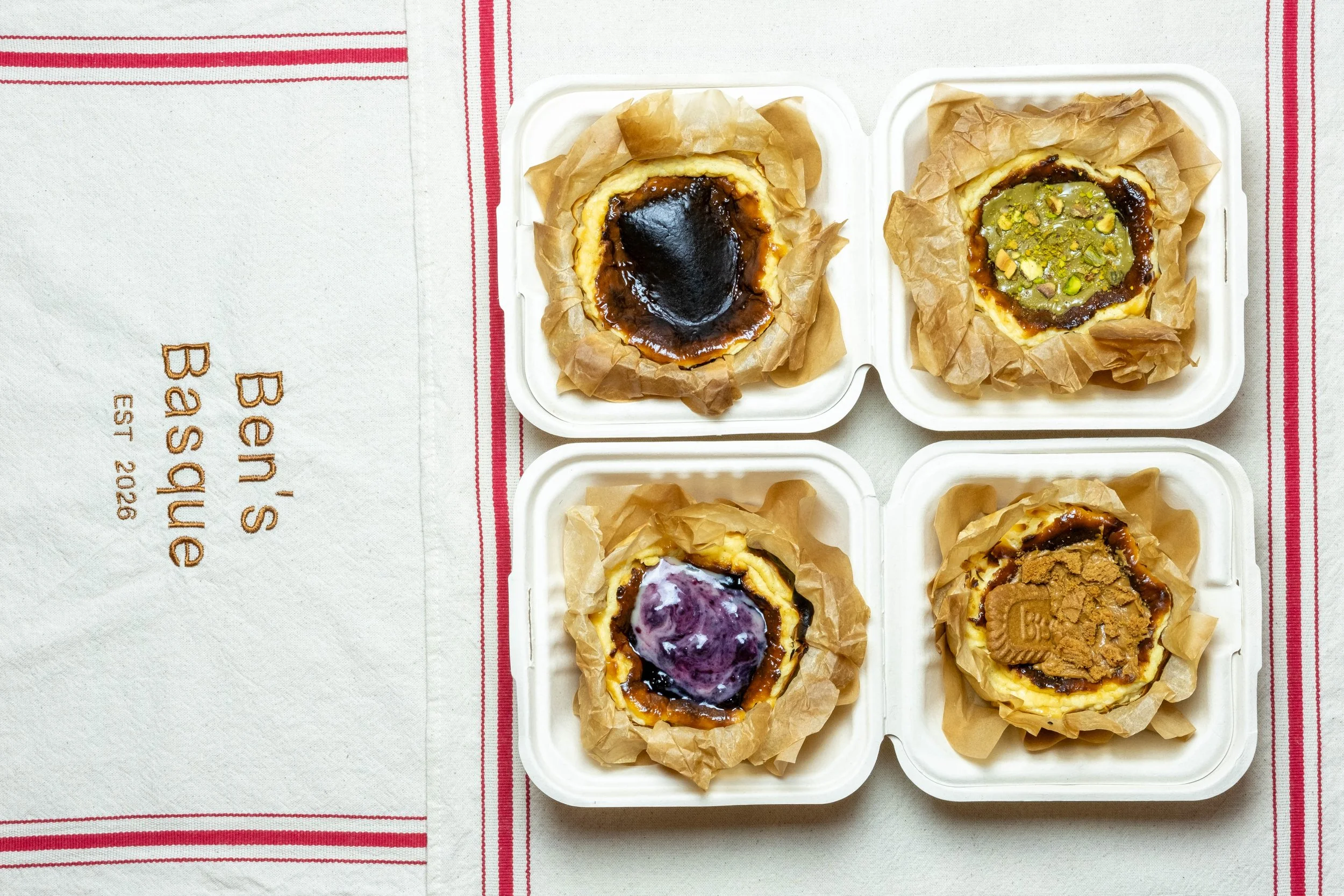 Four Basque cheesecakes topped with various toppings, placed on a linen cloth surface with red striped cloth underneath.