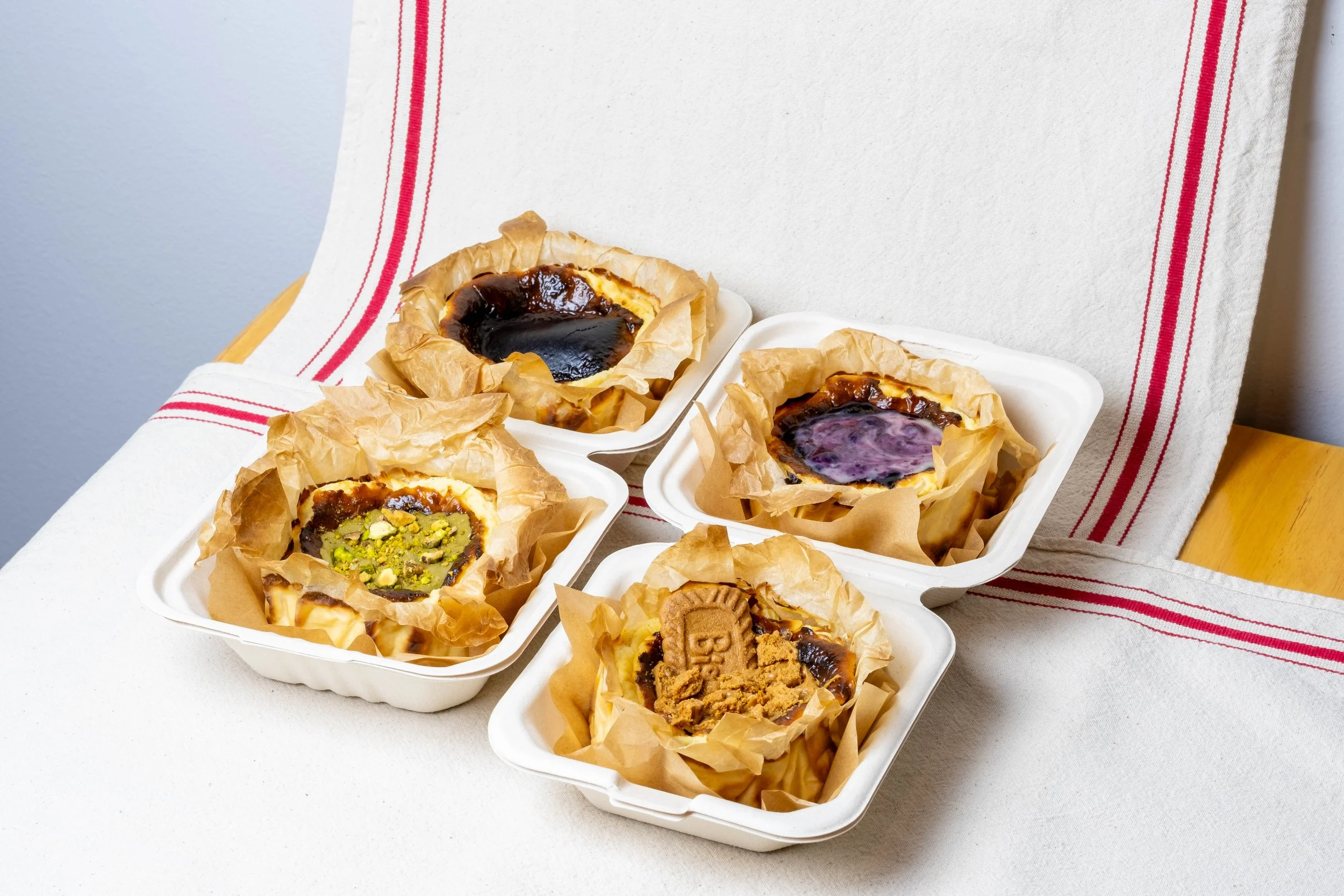 Four white containers with baked desserts on parchment paper, placed on a white textured tablecloth with red stripes, against a plain white wall.