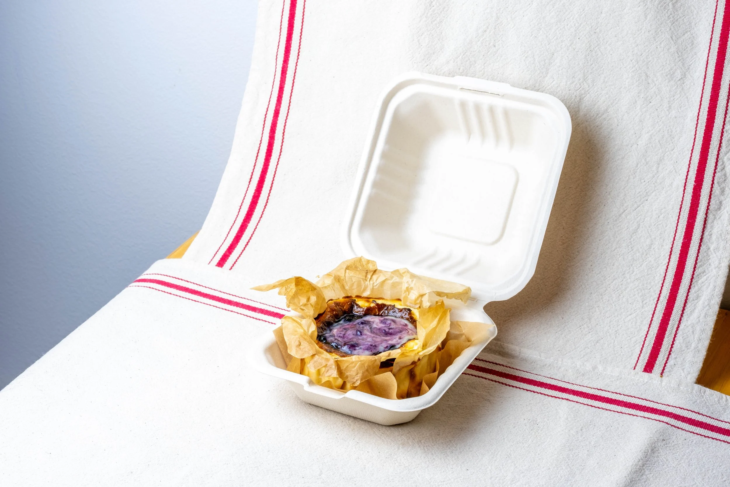 Bento Lemon Blueberry Basque cheesecake in a to go container, placed on a white table with a red-striped cloth.