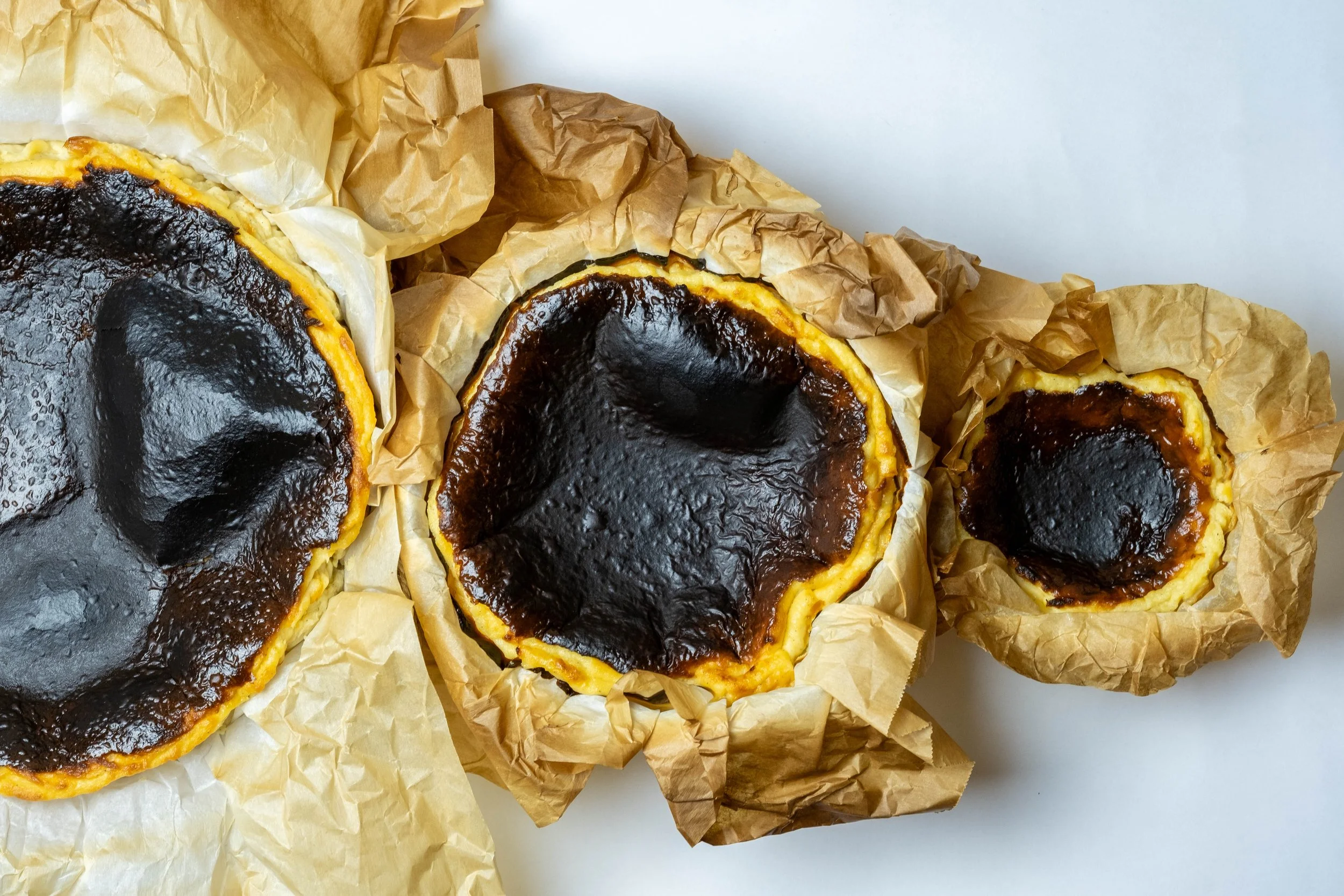 Three burnt cheesecakes on crumpled brown parchment paper on a white surface.