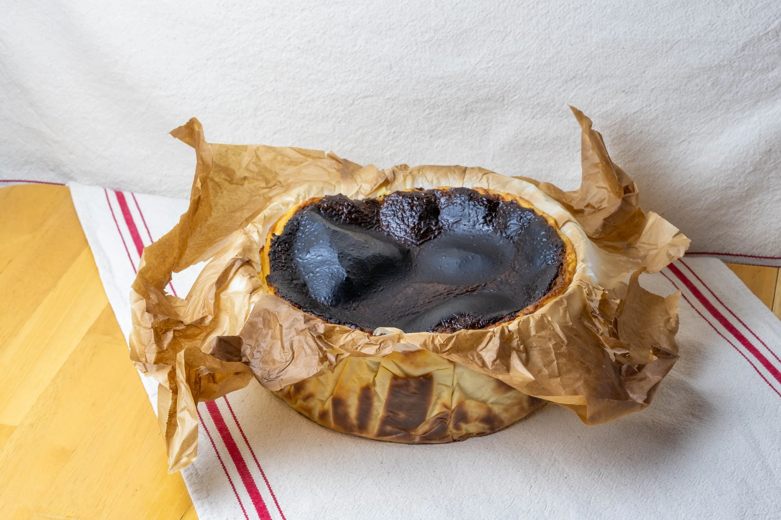 A chocolate tart with a shiny, dark chocolate glaze, nestled in brown parchment paper on a yellow wooden surface with a white cloth featuring red stripes.