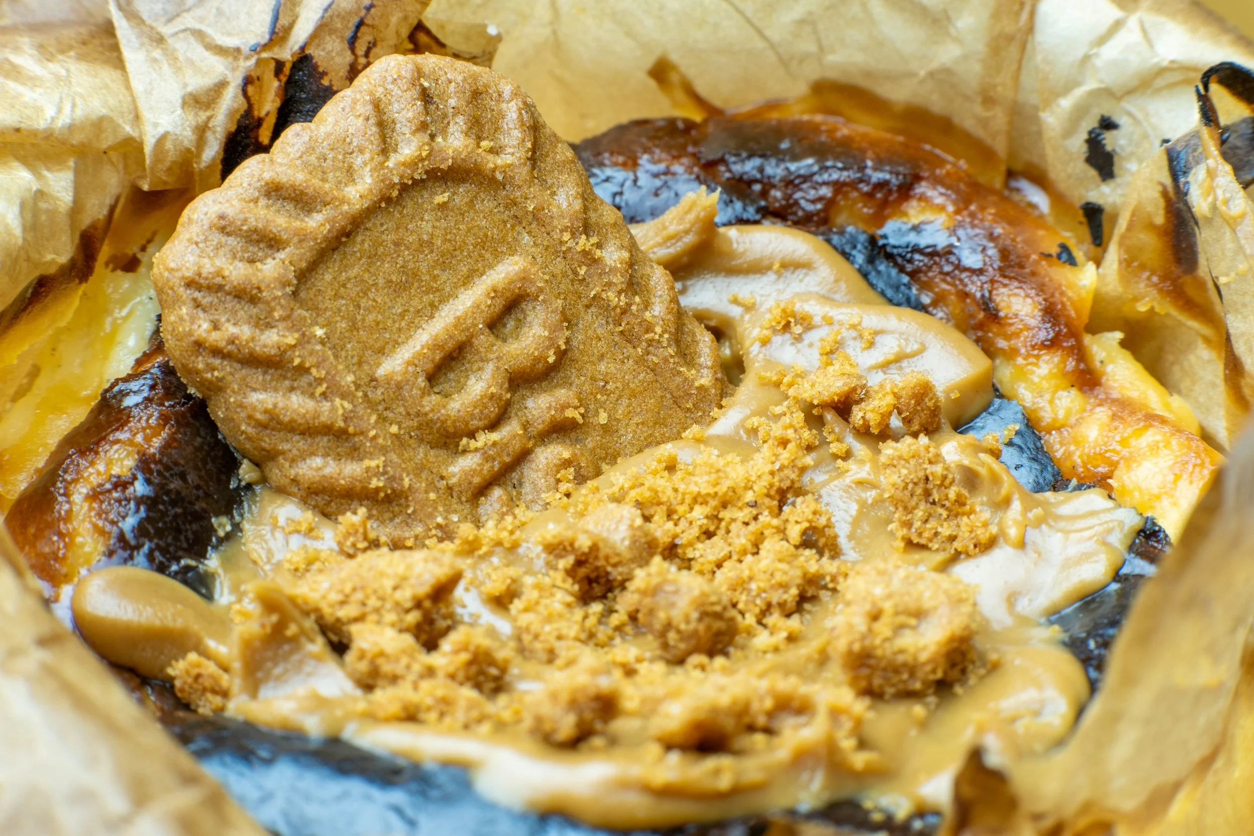 Close-up of a dessert with a large cookie with a stamped logo, caramel, chocolate, and crumbled toppings inside a paper wrapping.