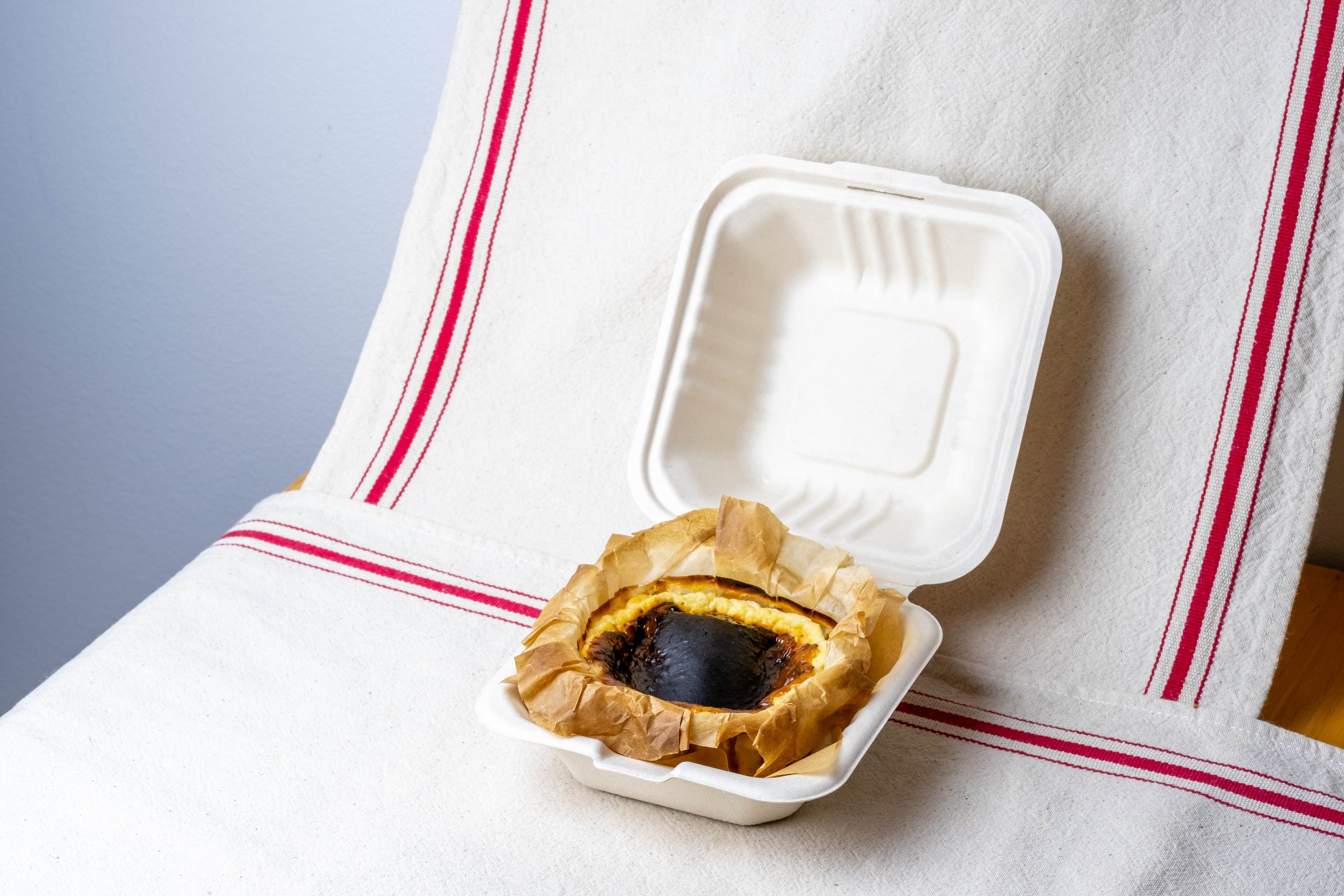 Bento Basque cheesecake in a to go container, placed on a white table with a red-striped cloth.