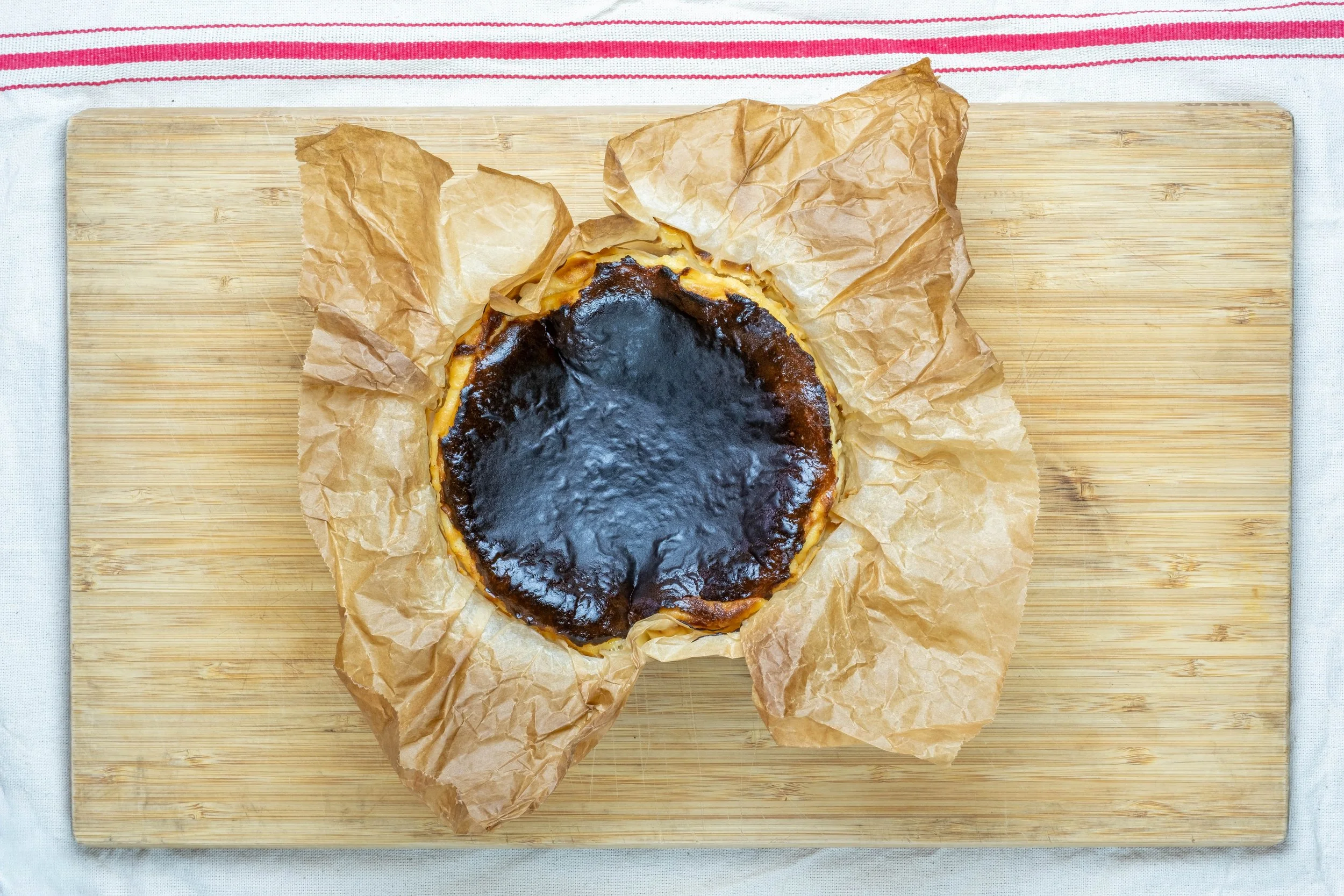 A burnt cheesecake with a dark caramelized top, on crumpled parchment paper, placed on a wooden cutting board.