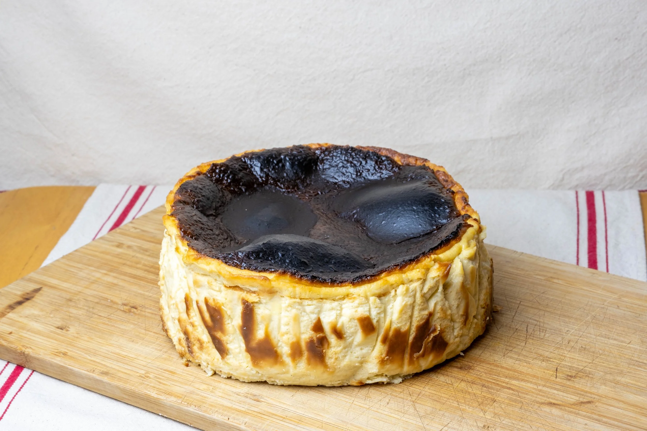 A baked cheesecake with a dark caramelized top on a wooden cutting board, with a white and red striped cloth underneath.