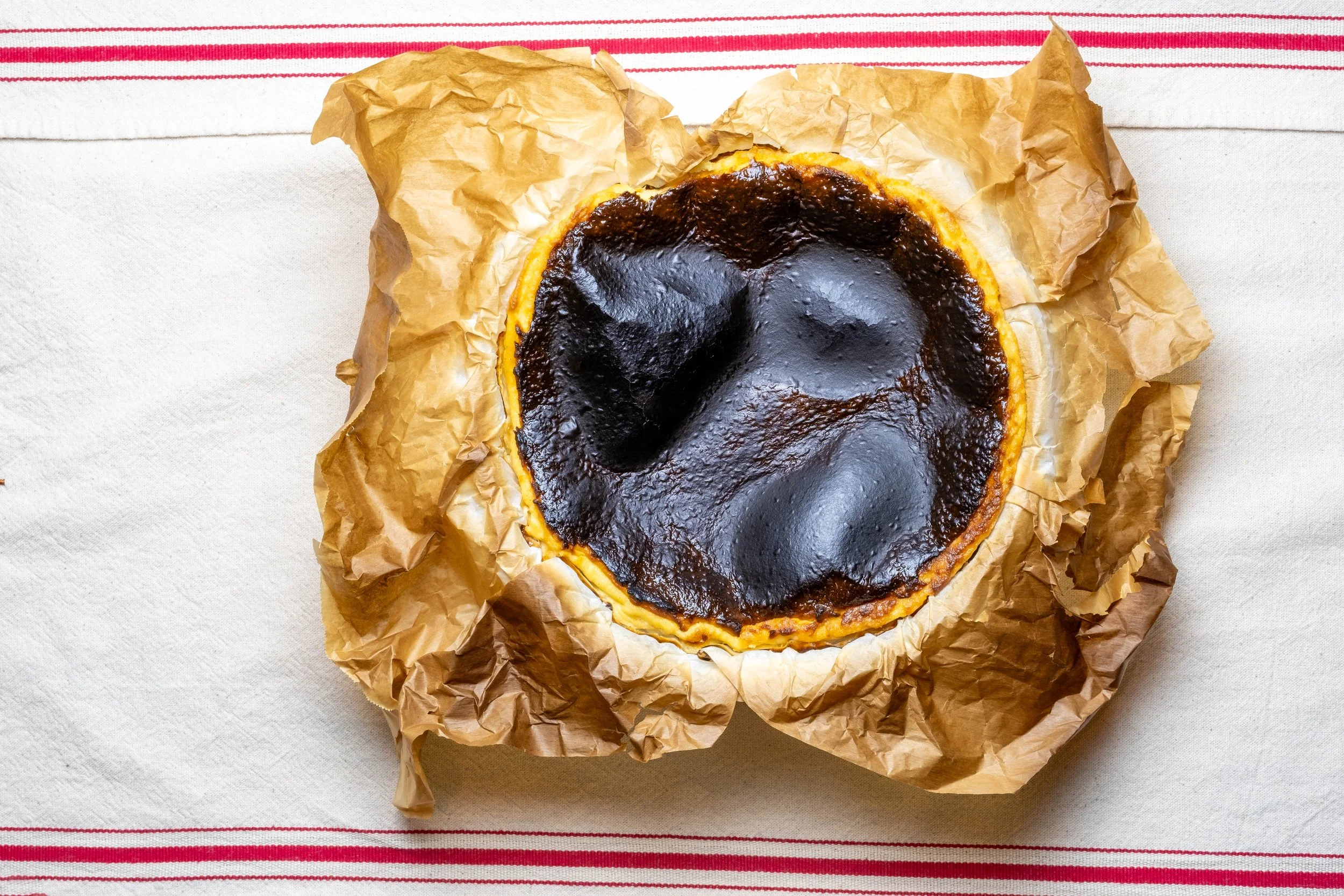 A burnt caramel cheesecake on crumpled parchment paper, placed on a white table with red striped cloth underneath.