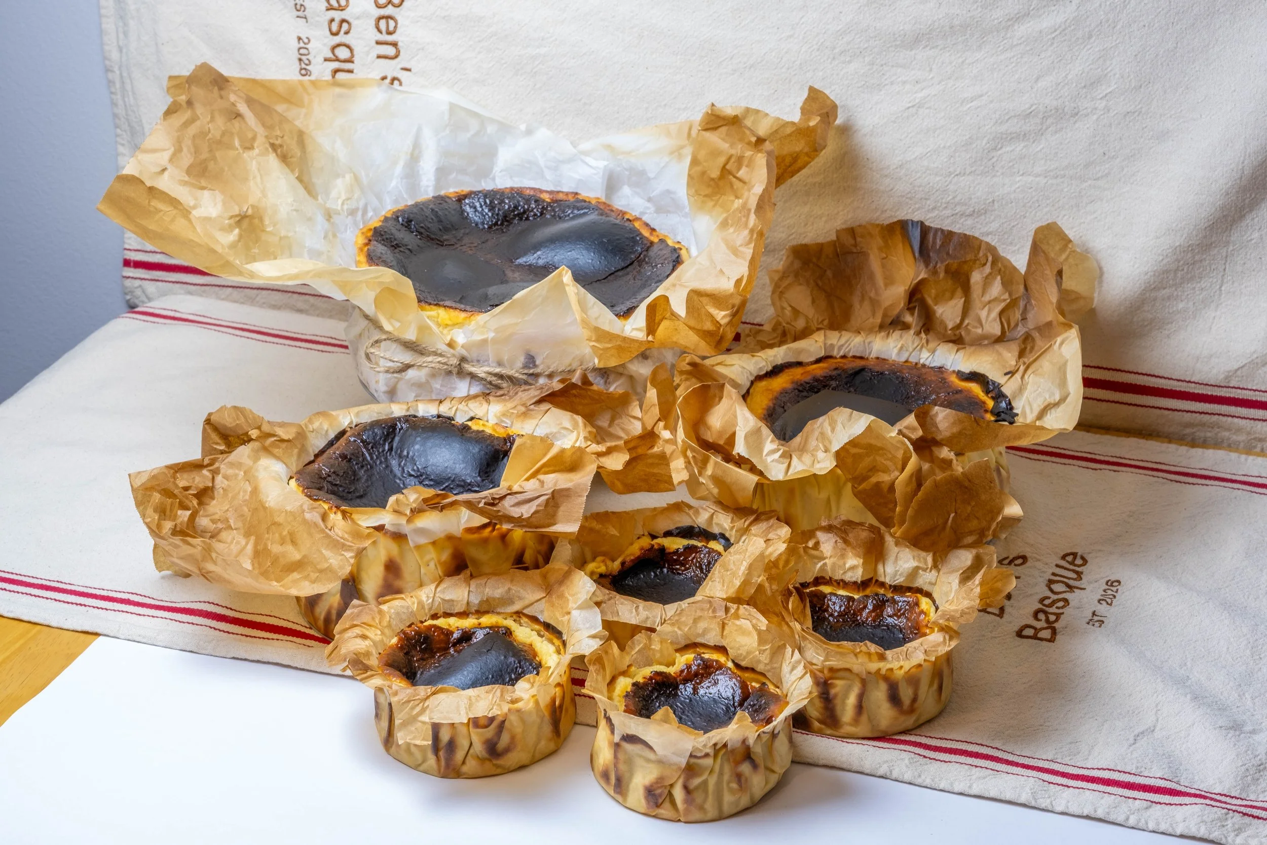 Multiple Basque cheesecakeswrapped in crinkled parchment paper, with burnt tops, placed on a white surface with a striped cloth in the background.