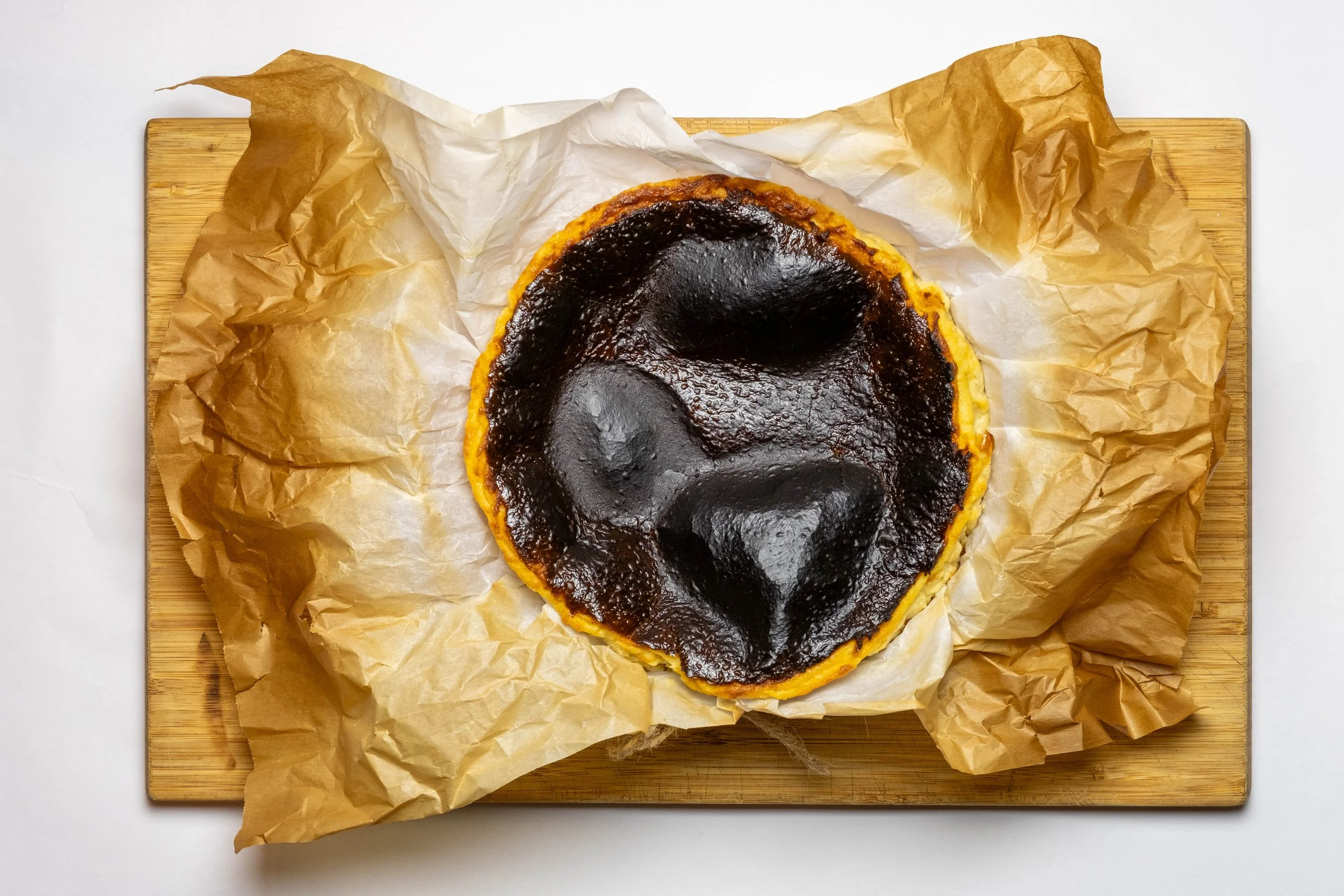 A burnt Basque cheesecakes  with a cracked, dark top is on crumpled parchment paper on a wooden board.