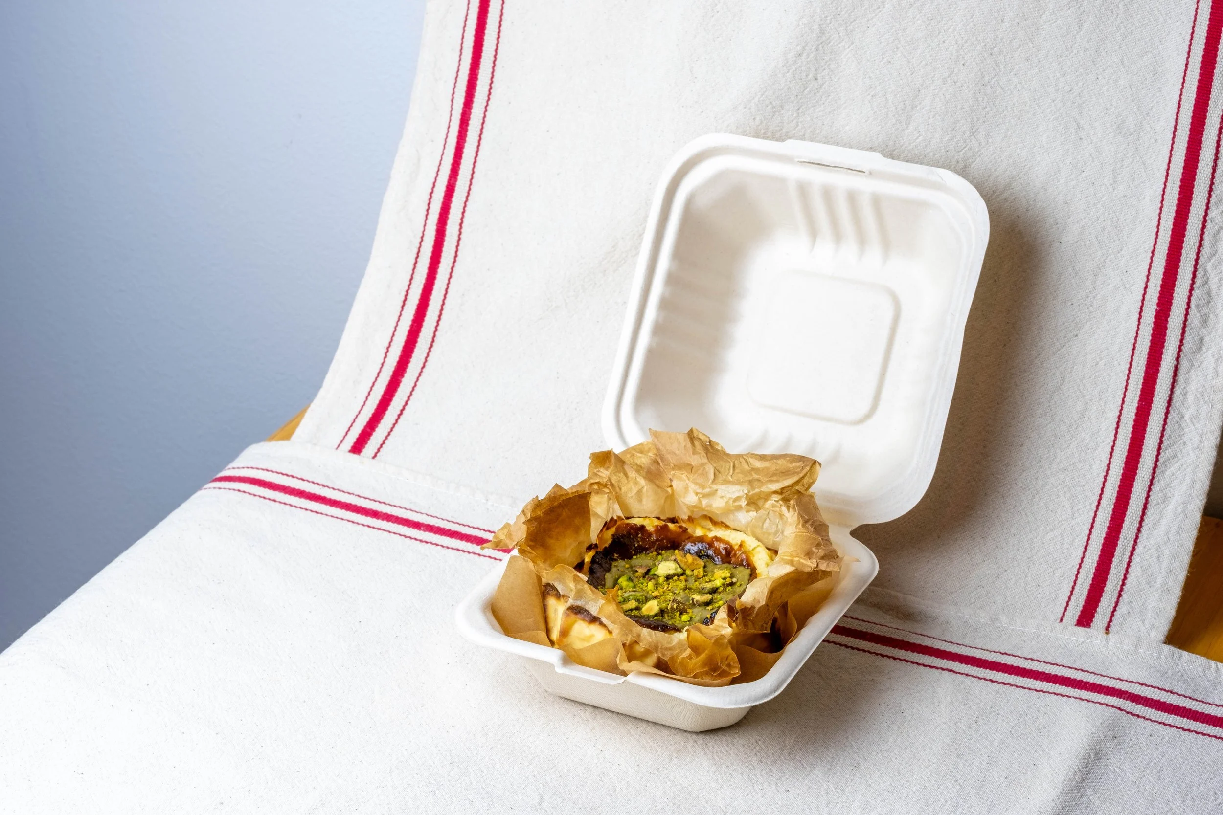 Bento Basque cheesecake with Pisti cream and pistachio topping in a to go container, placed on a white table with a red-striped cloth.