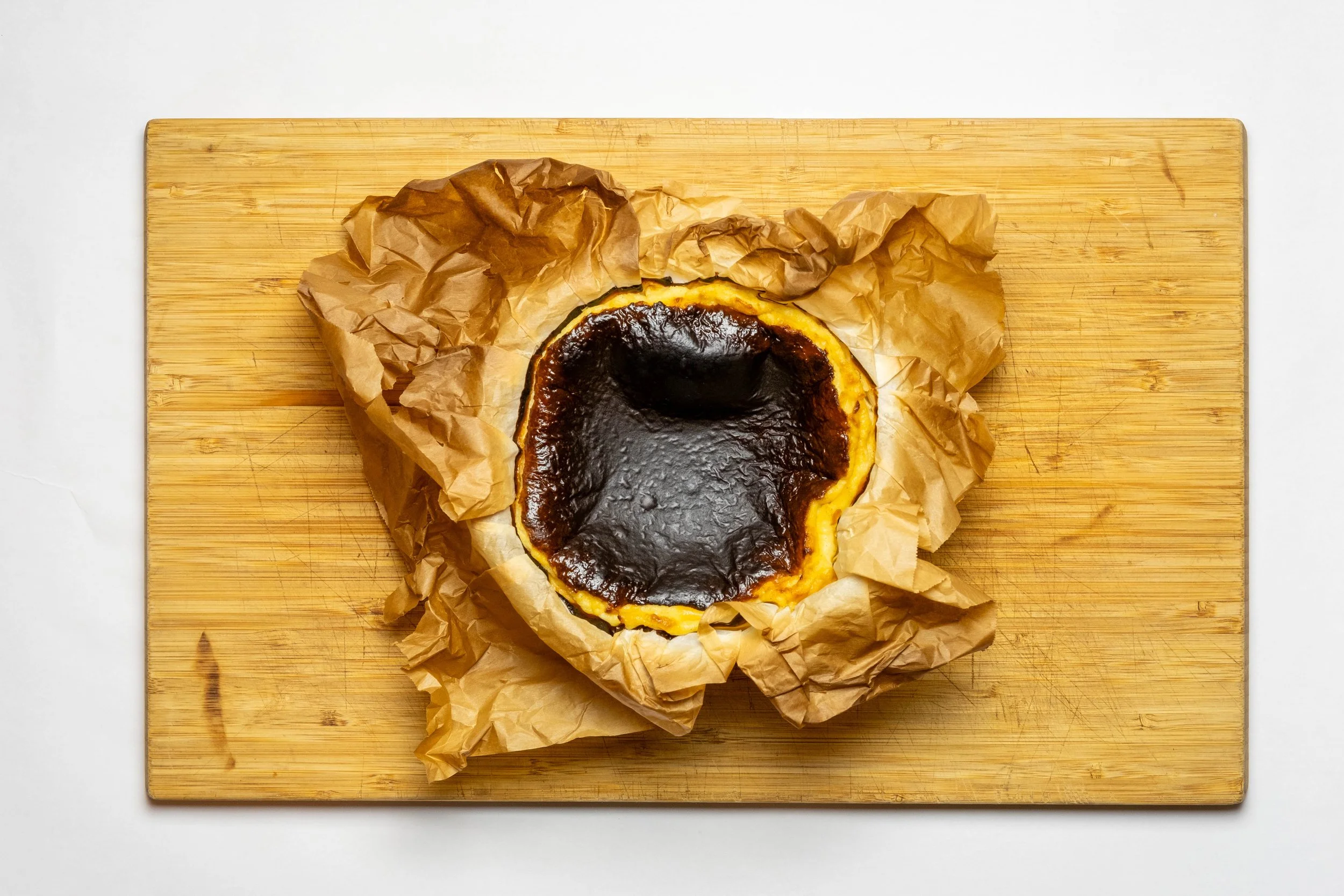 A burnt  Basque cheesecake with a dark, caramelized top sits on crumpled brown parchment paper on a wooden cutting board against a white background.