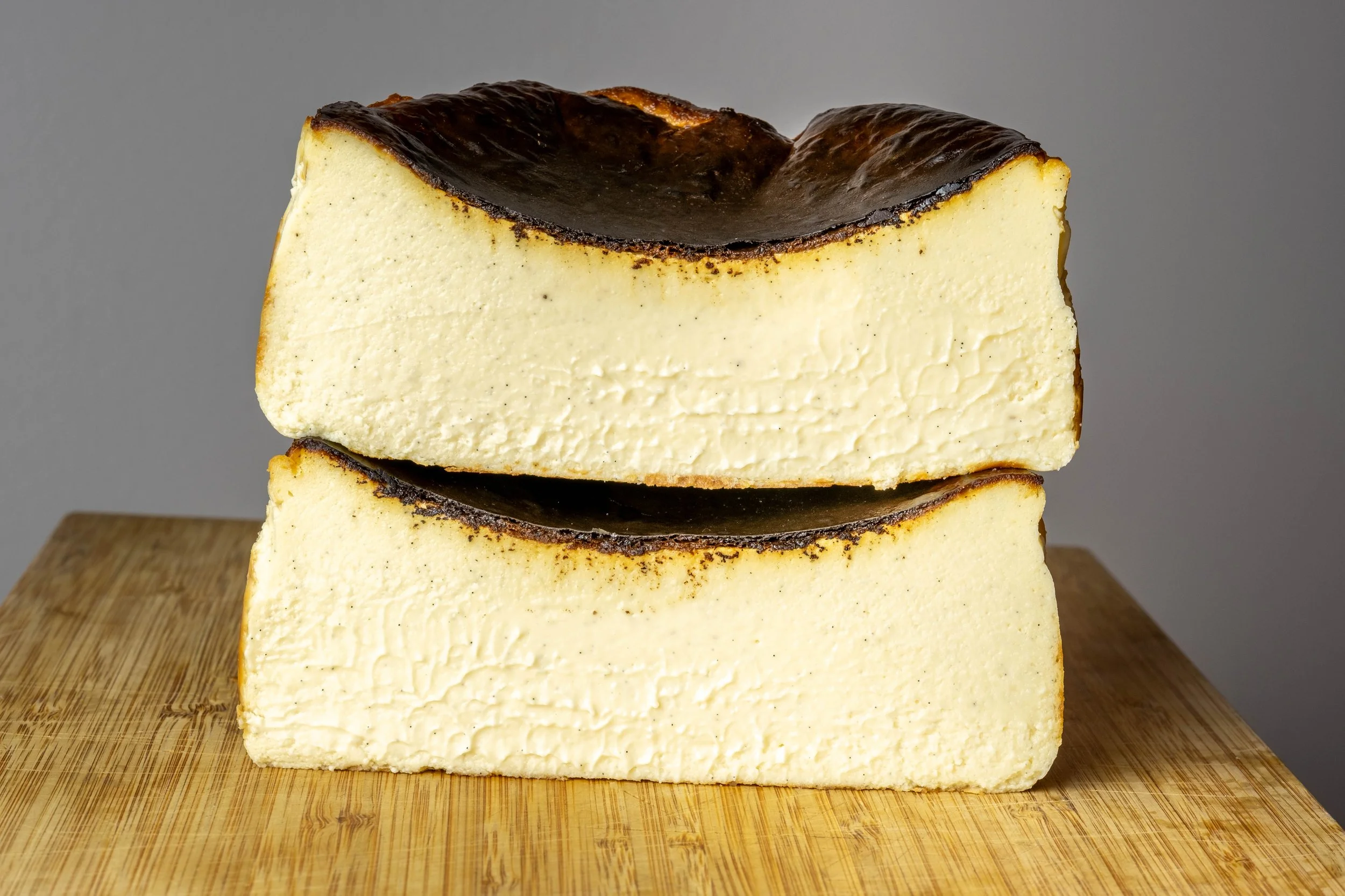 A close-up of a large block of cheesecake with a dark, burnt sugar crust on top, placed on a wooden cutting board against a plain gray background.