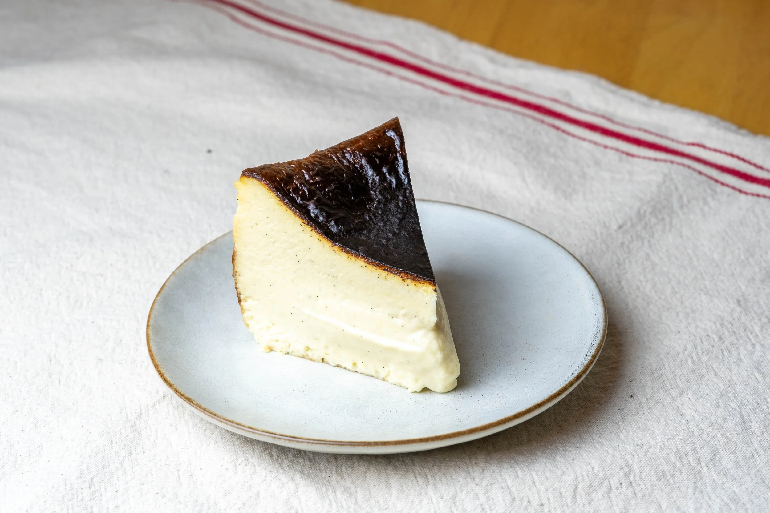 A slice of cheesecake with a dark caramelized top on a white plate with gold rim, placed on a light-colored fabric surface with a striped cloth in the background.