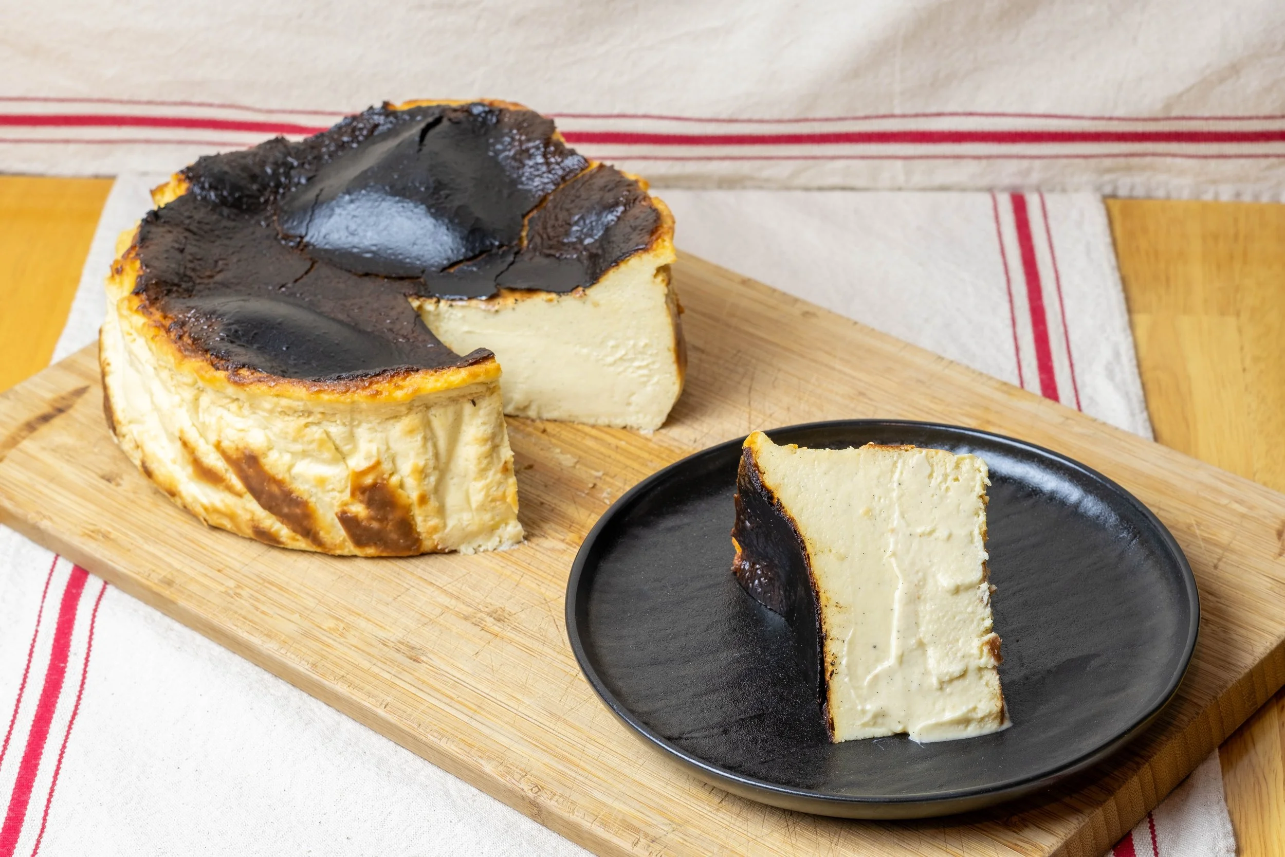 A slice of cheesecake with a chocolate topping on a black plate, placed on a wooden cutting board.