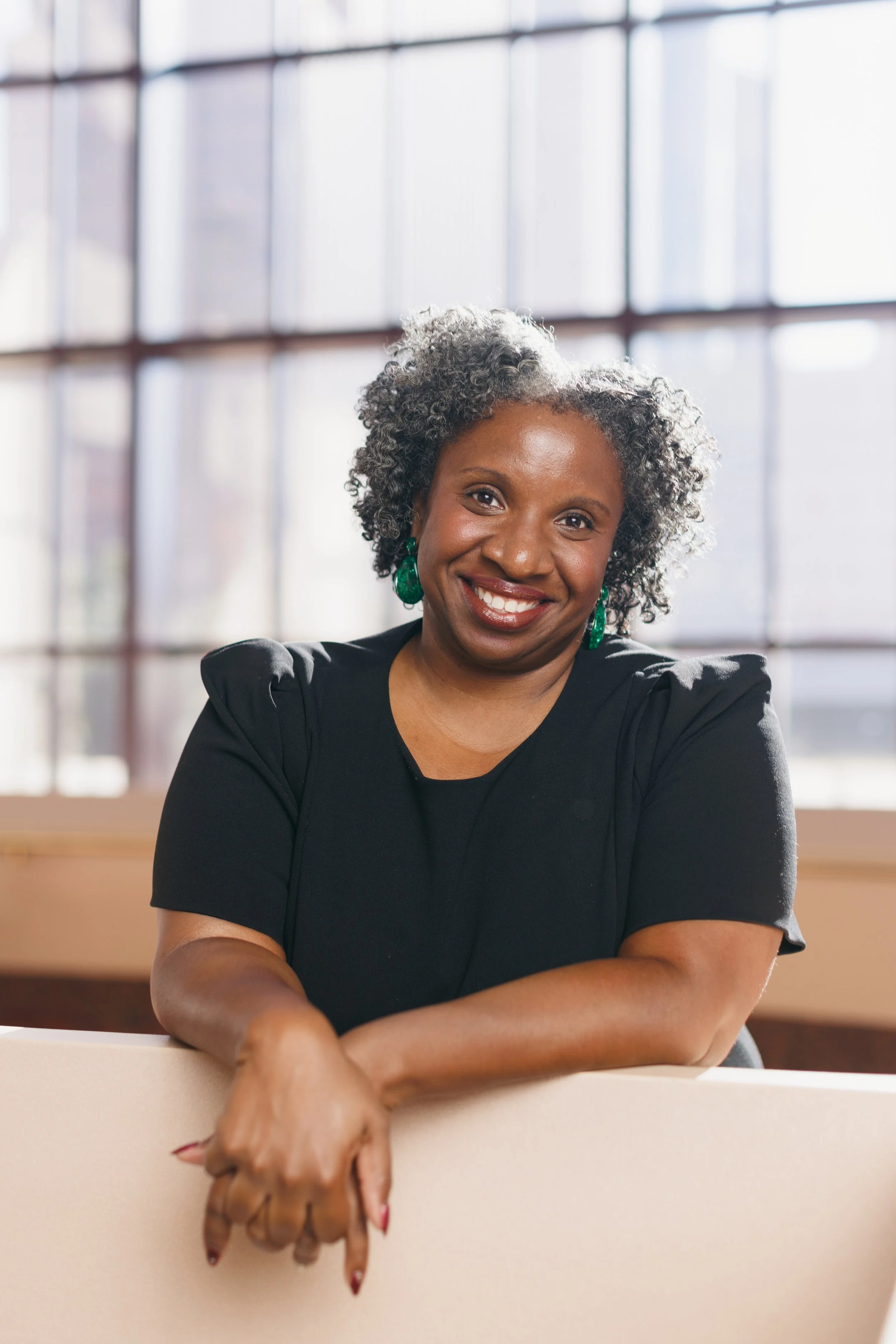 Smiling woman with short gray curly hair wearing green earrings, sitting at a table with folded arms, in front of a large window in the background letting in natural light.