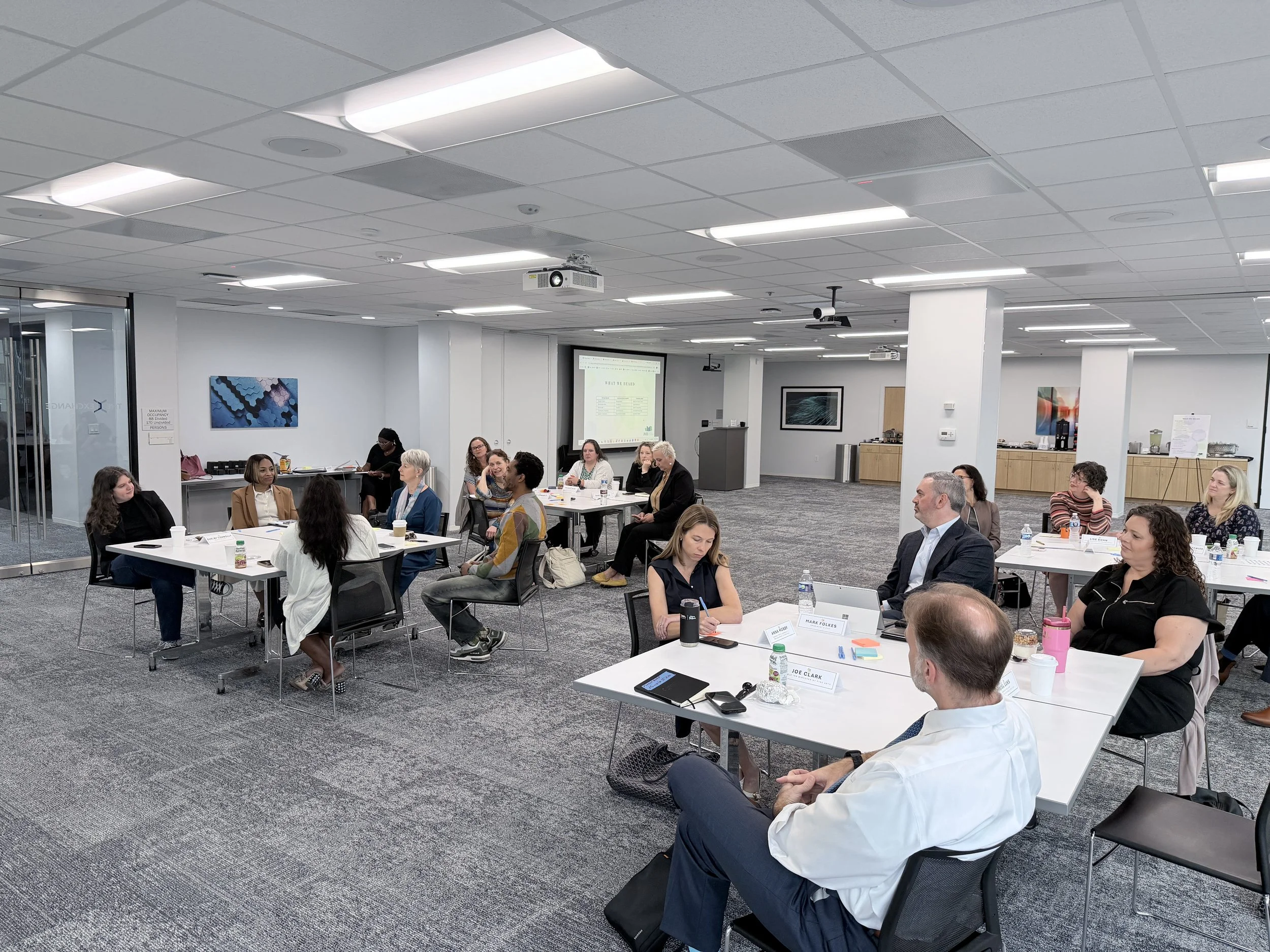 A diverse group of people attending a conference or seminar in a modern meeting room with white walls, artworks, and a large screen displaying a presentation.