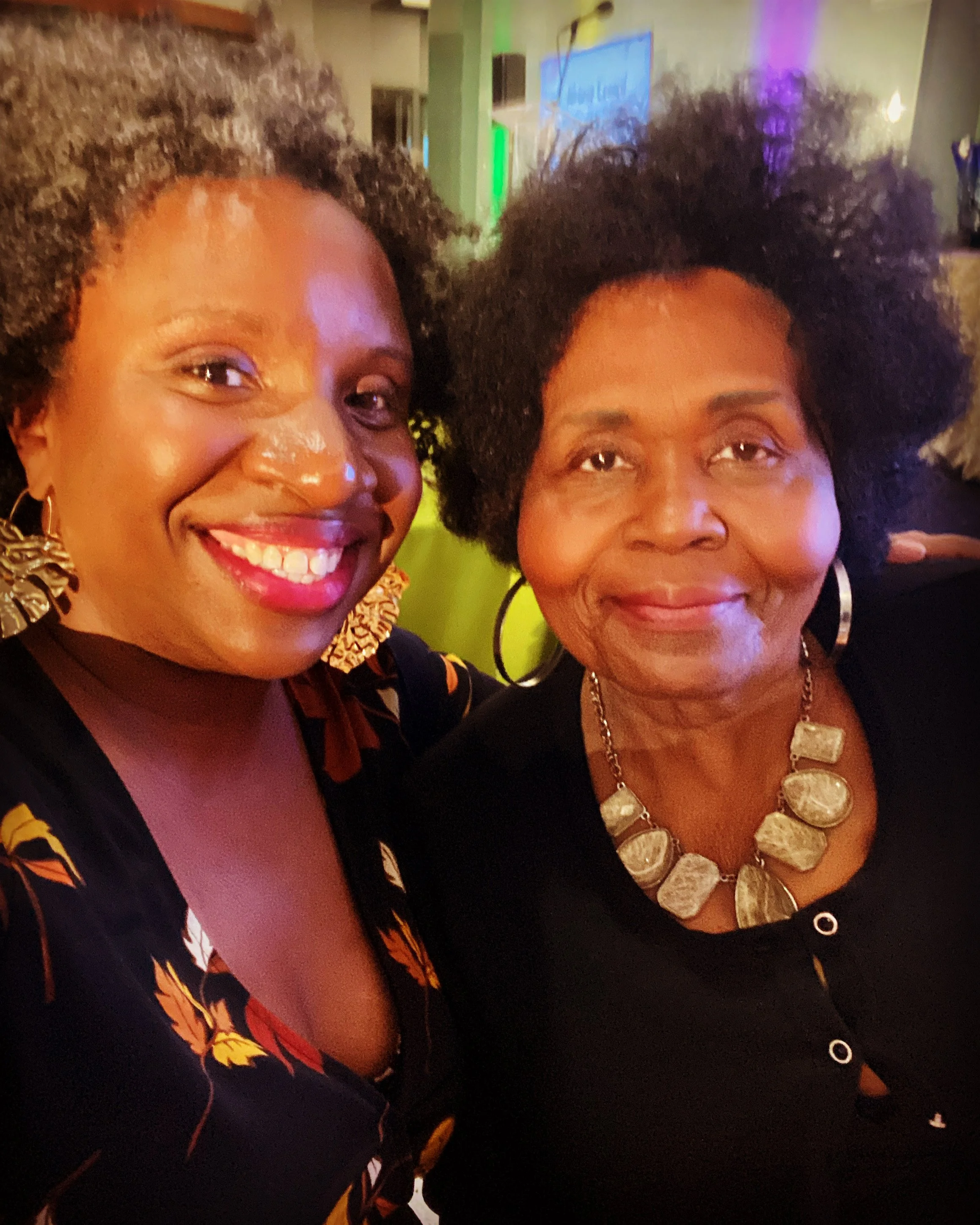 Two women smiling and posing for a photo in a dimly lit room. The woman on the left has curly hair, bright lipstick, and is wearing gold earrings and a patterned dress. The woman on the right has dark, voluminous hair, wears a large necklace with translucent stones and a black top with her hair styled in an afro.