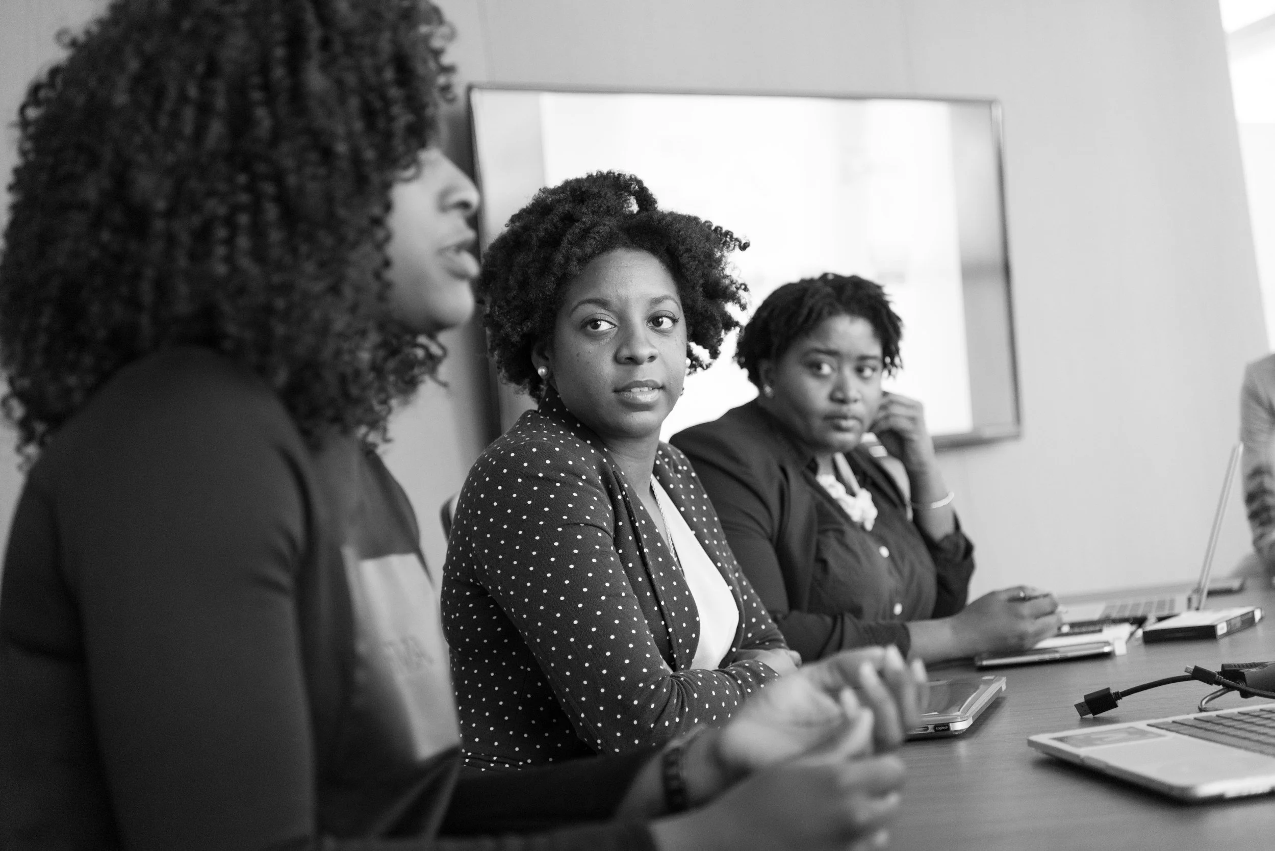 Three women sitting at a conference table during a meeting, with laptops and notebooks in front of them, engaged in discussion.