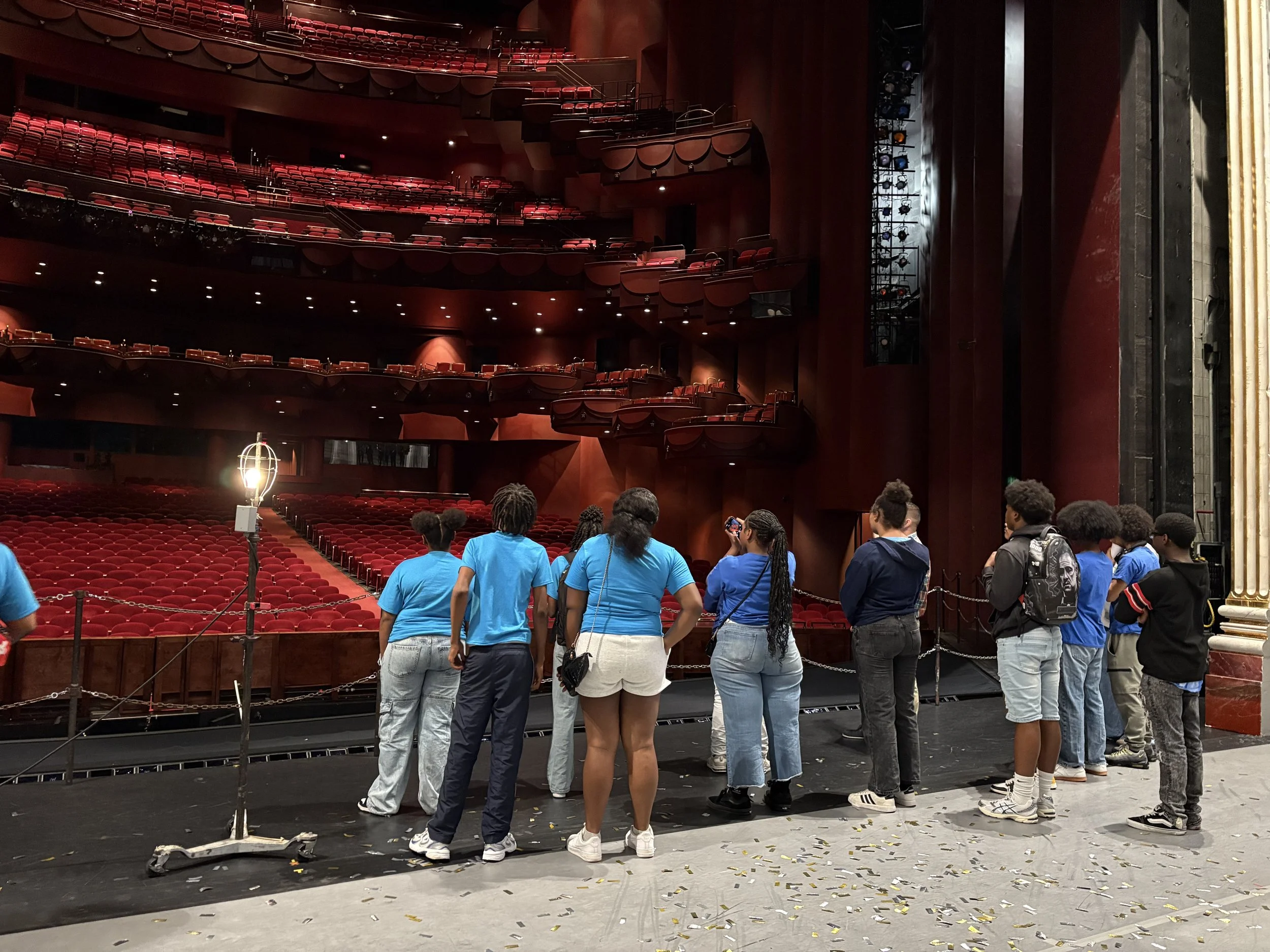 Young people in blue shirts standing on a stage in a theater, with red theater seats and stage lighting in the background.
