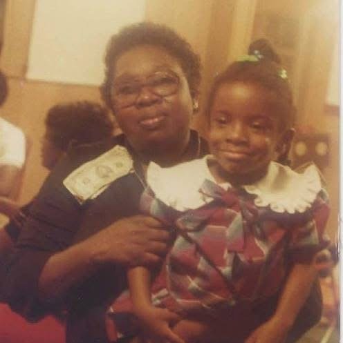 An adult woman hugging a young girl, both smiling at the camera, inside a warmly lit room.