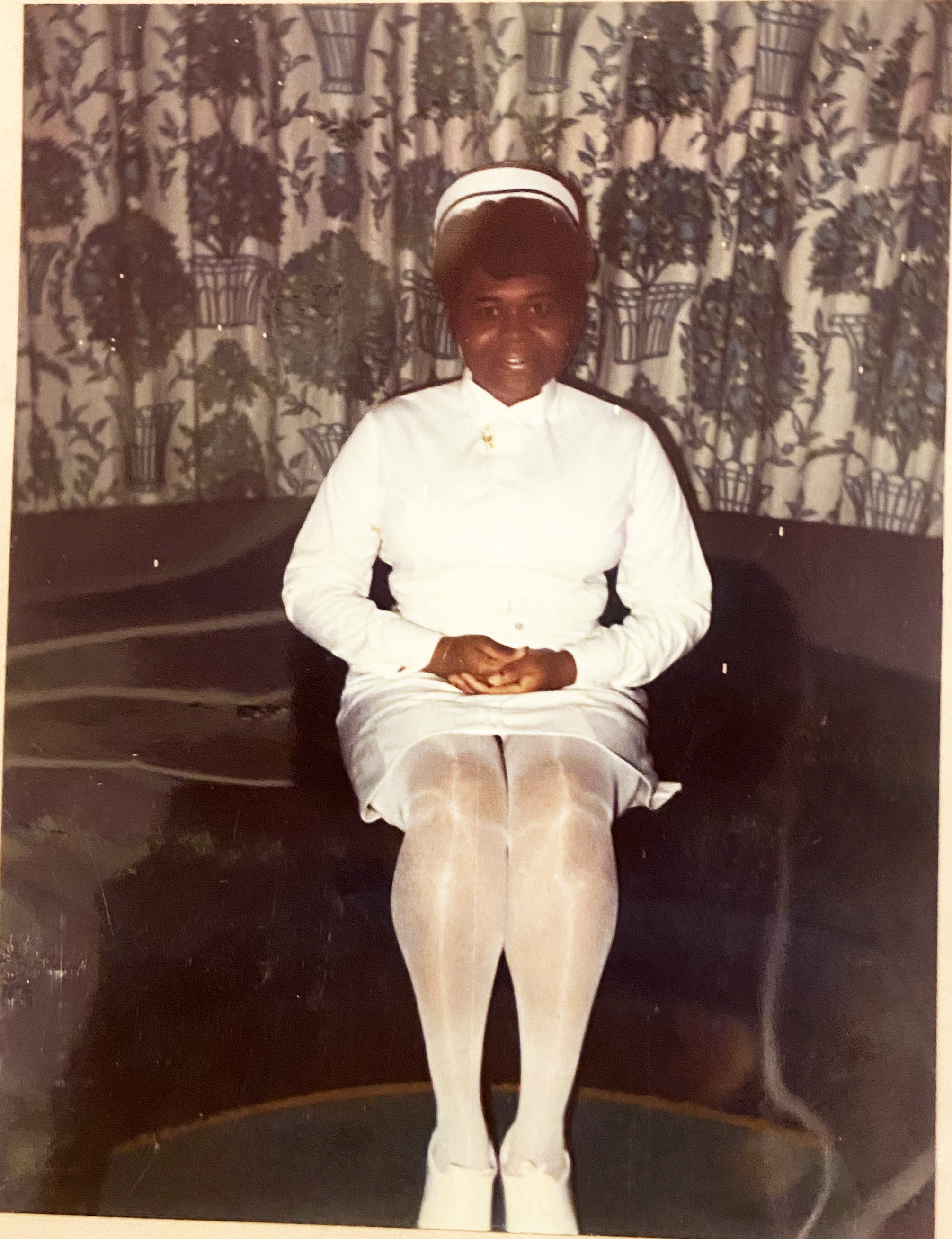 A woman with dark skin sitting in front of a floral-patterned curtain, wearing a white nurse uniform and white cap, with hands clasped in lap.